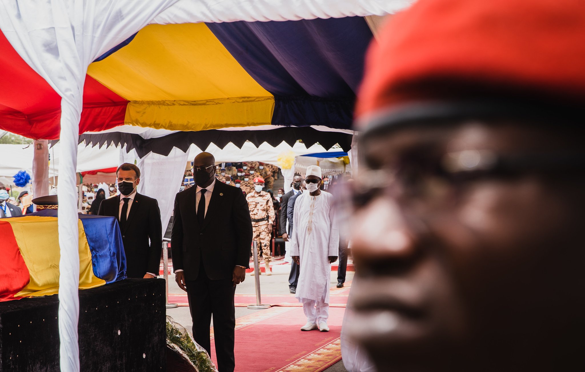 L'image montre une scène d'événement officiel, probablement une cérémonie. Au premier plan, il y a un homme portant des lunettes et un uniforme sombre, dont le visage est flou mais qui semble attentif. En arrière-plan, deux autres hommes, l'un en costume noir et l'autre en tenue traditionnelle blanche, marchent sous un chapiteau aux couleurs vives de jaune, rouge et bleu. Le chapiteau est décoré de tissu et crée une atmosphère formelle. La foule semble nombreuse et attentive, rendant l'ensemble de la scène solennelle et respectueuse. L'éclairage semble naturel, suggérant peut-être une belle journée ensoleillée.