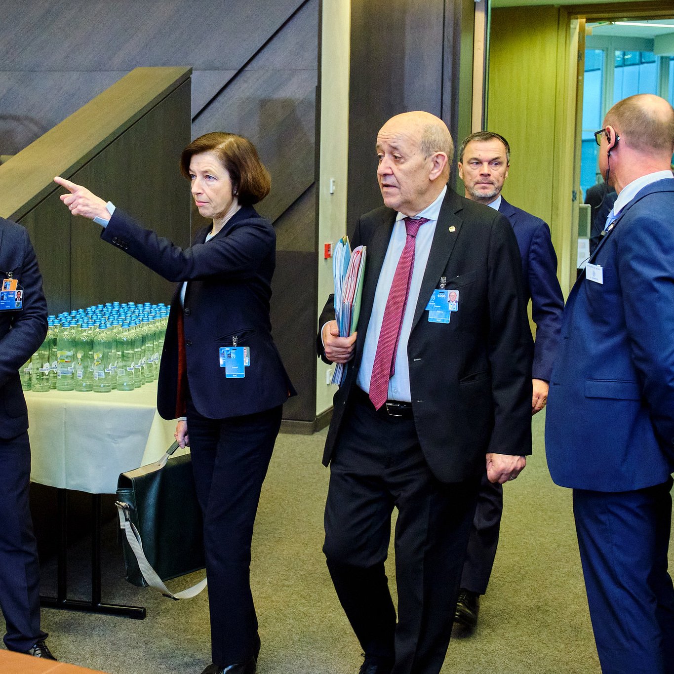 Dans cette scène, un groupe de personnes se déplace à l'intérieur d'un bâtiment moderne, probablement lors d'une réunion ou d'un événement officiel. Au premier plan, une femme, vêtue d'un costume professionnel et portant un document à la main, pointe dans une direction, semblant donner des indications. À ses côtés, un homme portant un masque de protection écoute attentivement, tandis qu'un autre homme, en costume noir, marche à ses côtés, apparemment engagé dans une conversation. En arrière-plan, d'autres participants sont visibles, suggérant une atmosphère animée et collaborative. Des bouteilles d'eau sont disposées sur une table à proximité, signalant la durée de l'événement. Le décor est contemporain, avec des murs en bois et des éléments de design épuré.
