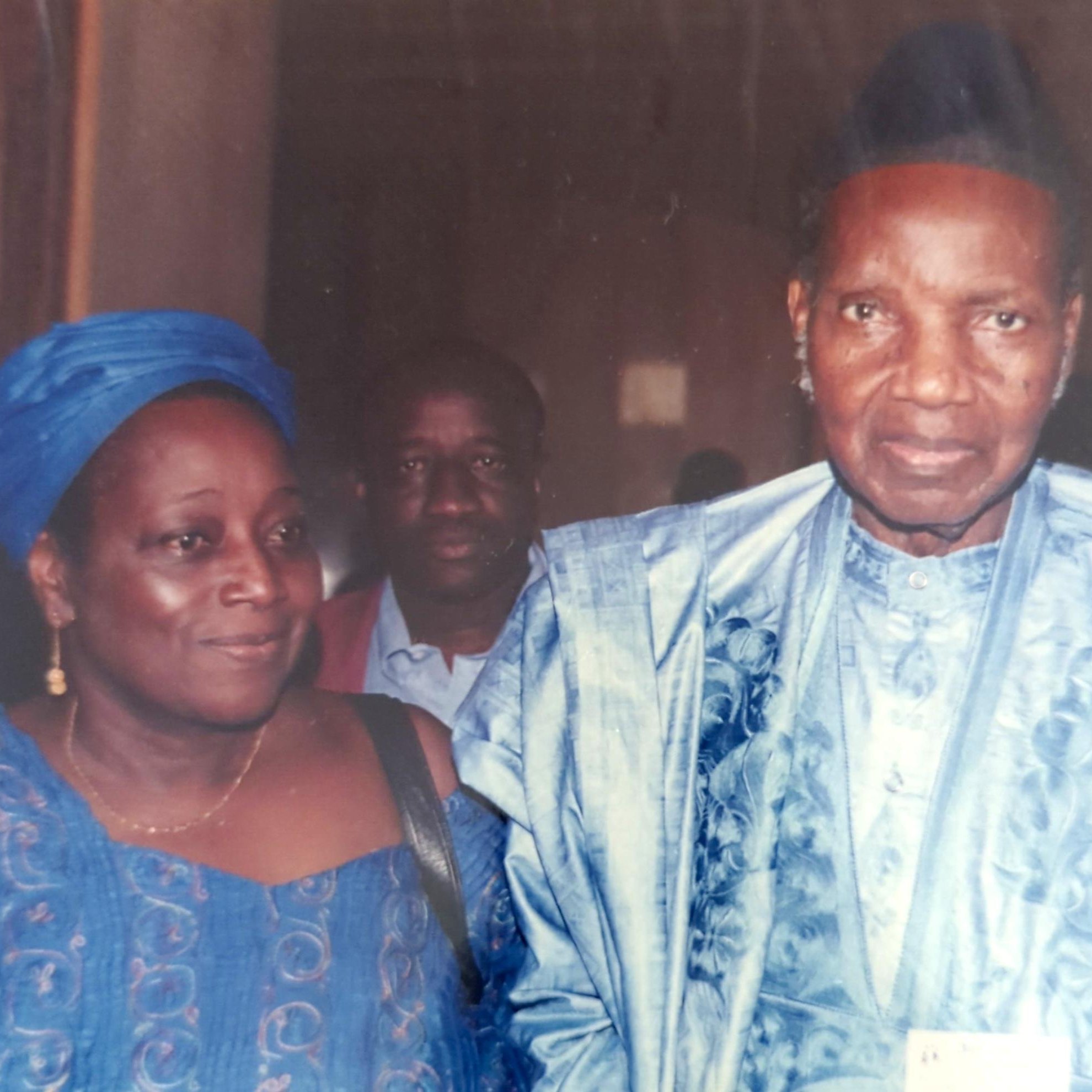 L'image montre trois personnes. À l'avant, un homme et une femme sont vêtus de costumes traditionnels, probablement en tissus d'importance culturelle. L'homme porte un chapeau et a un regard sérieux, tandis que la femme a un sourire et semble détendue. En arrière-plan, un troisième homme est visible, bien qu'il soit moins en avant-plan. L'ambiance de l'image semble formelle.