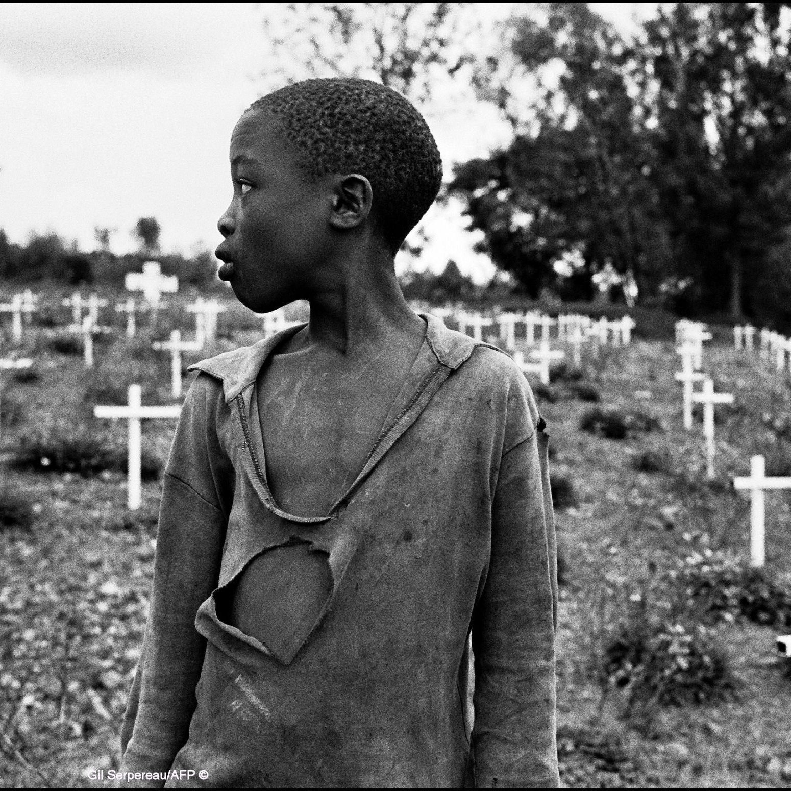 L'image présente un jeune garçon qui se tient dans un cimetière parsemé de croix blanches, symbolisant des tombes. Son regard est tourné vers la gauche, et on peut percevoir une expression de réflexion ou de tristesse sur son visage. Il porte un vêtement déchiré, soulignant une certaine vulnérabilité ou pauvreté. En arrière-plan, le paysage est entouré d'arbres, créant une atmosphère de mélancolie. Les croix blanches, disposées sur la pente, évoquent la mémoire et le souvenir de ceux qui ont perdu la vie.