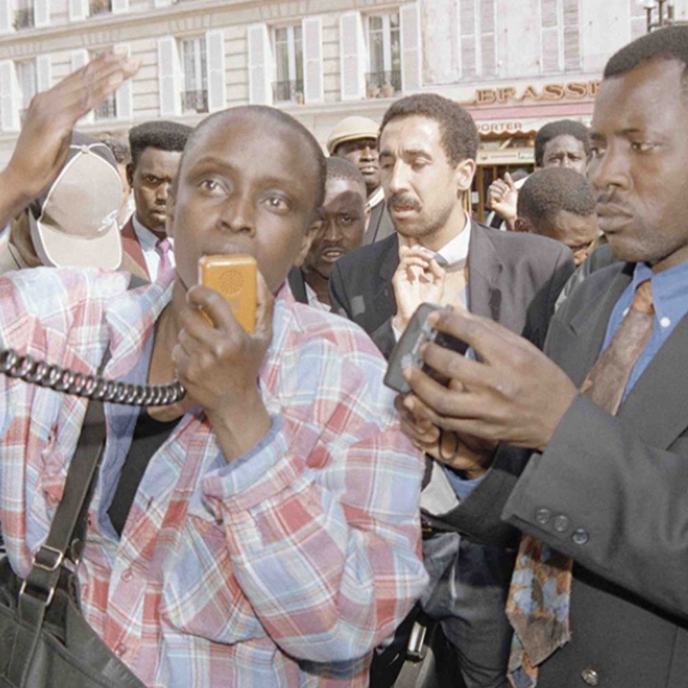 L'image montre un groupe de personnes rassemblées dans un espace public animé. Au premier plan, deux hommes attirent l'attention. L'un d'eux, portant une chemise à carreaux et tenant un microphone, semble s'exprimer avec passion, les bras levés, signalant une forte émotion. L'autre homme, en costume sombre, tient un appareil photo, observant et capturant le moment. En arrière-plan, on peut apercevoir une foule diverse, des visages attentifs, écoutant ou réagissant à la situation. L'atmosphère générale est dynamique, avec un sentiment de mobilisation ou de protestation.