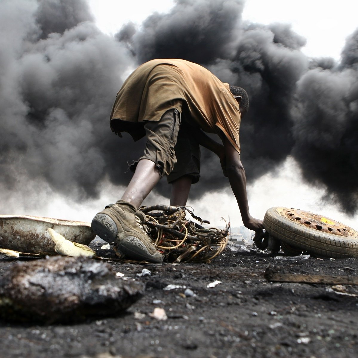 L'image montre un environnement très sombre et pollué, avec d'épaisses colonnes de fumée noire s'élevant dans l'air. Au premier plan, un jeune homme est accroupi, en train de manipuler des débris. Il porte des vêtements simples, à moitié sales, et semble concentré sur ce qu'il fait. Son environnement est chaotique, avec des morceaux de métal, de la ferraille et des pneus éparpillés autour de lui. La scène dégage une atmosphère de difficulté et de lutte, illustrant les conditions de vie dans cet endroit touché par la pollution et la dégradation environnementale.