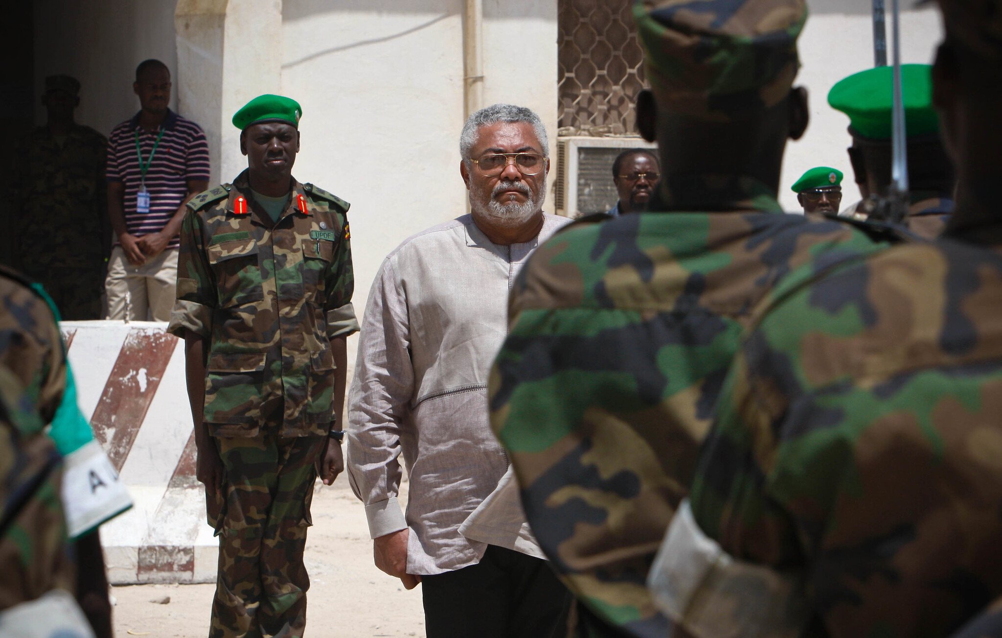 L'image montre un homme d'âge moyen au centre, avec des cheveux poivre et sel et une barbe grise. Il porte une chemise claire. Il semble sérieux et concentré, entouré de militaires en uniforme camouflage. Ces soldats, qui se tiennent en rangs, regardent vers lui. En arrière-plan, on peut voir un bâtiment aux murs clairs et un voisinage qui évoque un environnement militaire. L'atmosphère est formelle, soulignant l'importance de la scène.