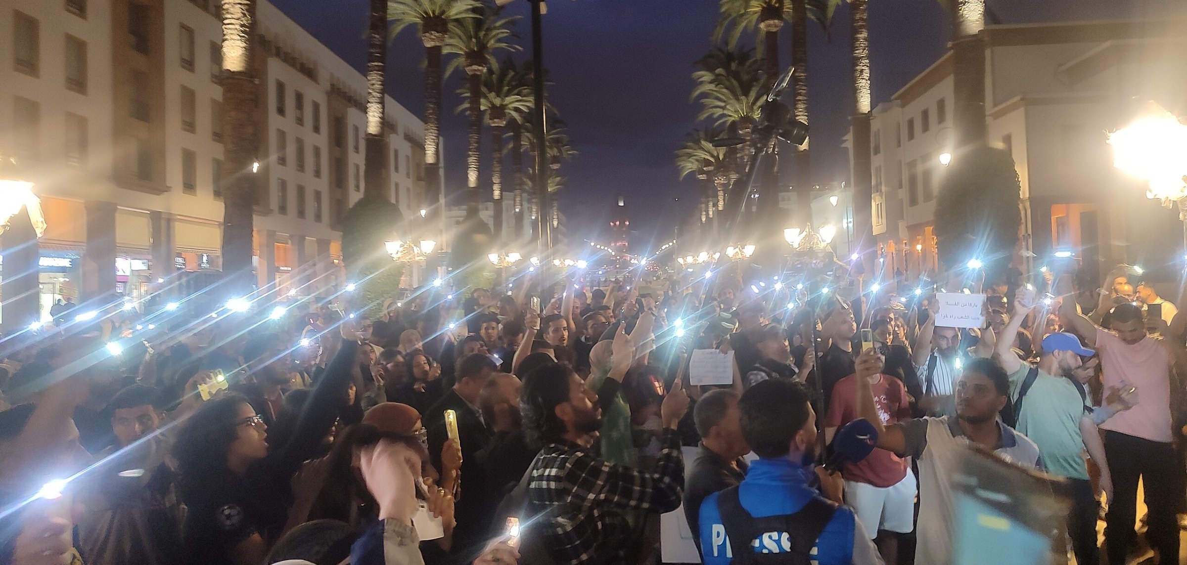 L'image montre une grande foule rassemblée dans une rue animée, éclairée par des lampadaires et entourée de palmiers. La scène se déroule en soirée, avec un ciel crépusculaire. Beaucoup de personnes tiennent des téléphones ou des lumières, créant un effet lumineux impressionnant. Les gens semblent mobilisés, certains tenant des pancartes et s'exprimant avec passion. L'atmosphère est énergique et engagée, reflétant une ambiance de manifestation ou d'événement public où les participants cherchent à faire entendre leur voix.