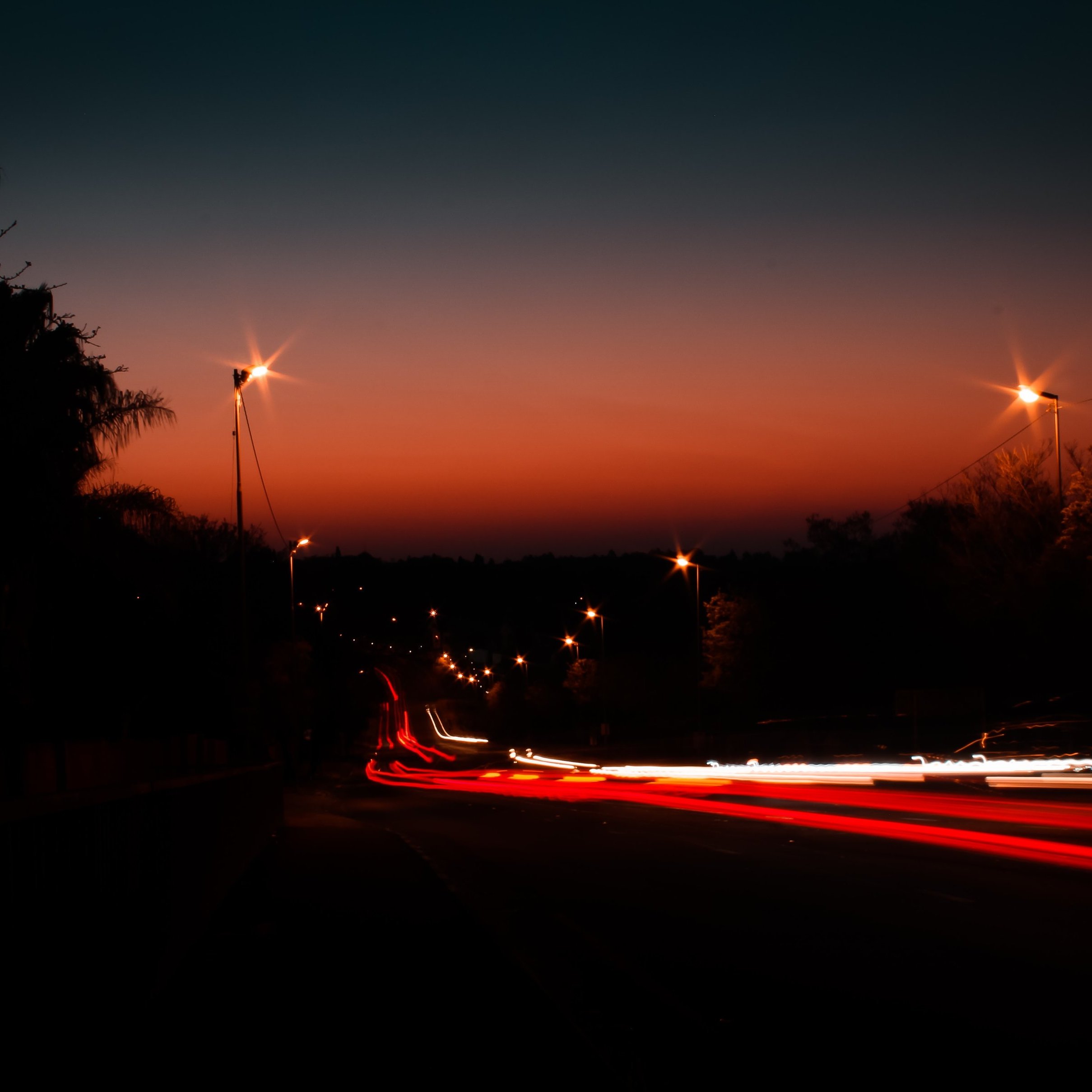 Imagine une scène tranquille à l'heure où le jour se transforme en nuit. Le ciel se teinte de dégradés d'orange et de bleu sombre, créant une ambiance douce et apaisante. Au loin, on aperçoit une route bordée d'arbres, presque silhouettes noires se découpant à l'horizon. Des lampadaires illuminent le chemin, leurs lumières jaunies ajoutant une touche chaleureuse à la scène. Des traces de lumière, comme des lignes fluides et vives, suggèrent des voitures qui passent rapidement, laissant derrière elles des filaments rouges et blancs. L'ensemble crée une atmosphère calme, invitant à la contemplation de ce moment transitionnel entre le jour et la nuit.
