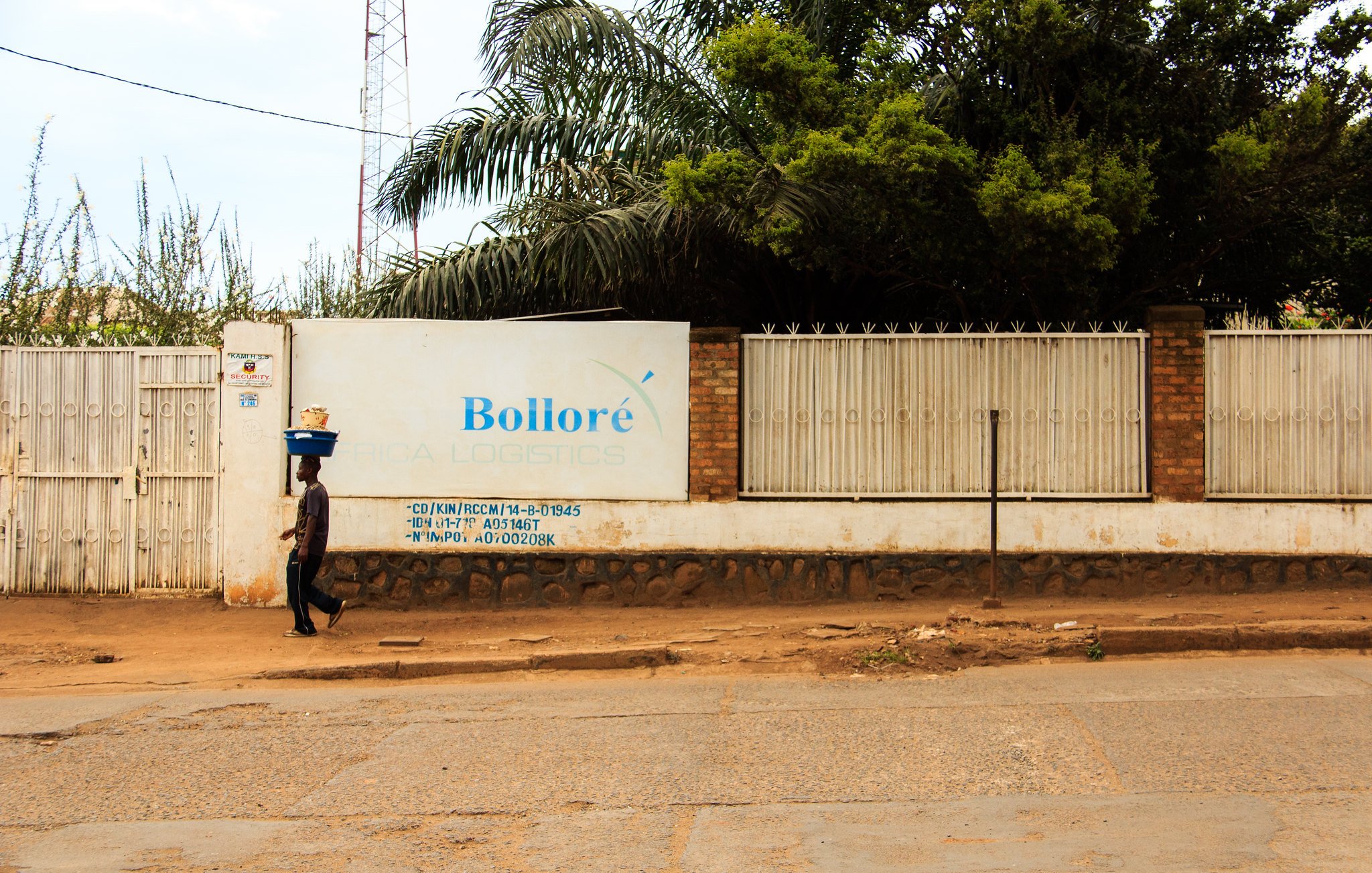 L'image montre un paysage urbain avec une route en terre battue. Sur le côté gauche, une personne marchant lentement porte un fardeau sur sa tête, ce qui donne une impression de travail acharné. À droite, un grand mur blanc affiche le nom "Bolloré" ainsi que des inscriptions et des informations liées à l'entreprise, à côté d'une clôture en métal. En arrière-plan, quelques arbres et une tour de communication sont visibles, ajoutant une touche de verdure à la scène. L'atmosphère semble calme, avec un éclairage naturel qui suggère une journée ensoleillée.
