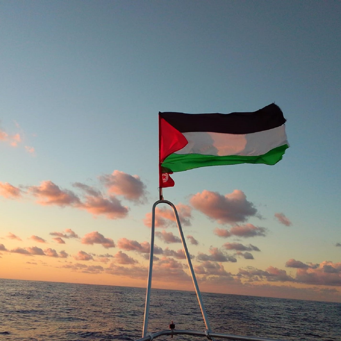 L'image montre un drapeau palestinien flottant sur un mât, avec un vaste horizon marin au fond. Le ciel est teinté de couleurs chaudes, probablement à l'aube ou au crépuscule, avec des nuages épars. Les couleurs du drapeau sont distinctes : le noir, le blanc, le vert et le rouge. La mer est calme et reflète doucement les teintes du ciel, créant une ambiance paisible et solennelle.