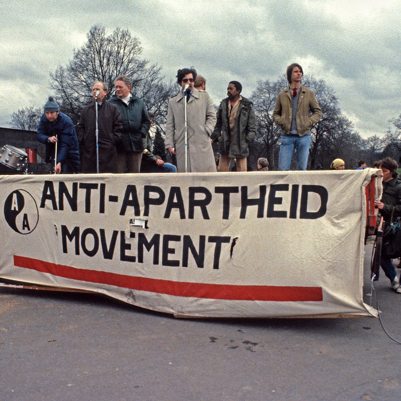 Cette image représente un rassemblement pour le mouvement anti-apartheid. On voit une scène sur un camion, avec plusieurs personnes qui parlent à un microphone. La bannière au premier plan affiche le texte "ANTI-APARTHEID MOVEMENT" en lettres majuscules, souligné par une bande rouge. L'environnement est urbain, avec des arbres dénudés en arrière-plan et un ciel nuageux, ce qui crée une atmosphère sérieuse et engagée. Les participants semblent passionnés, témoignant de l'importance de la lutte pour l'égalité et les droits civiques.