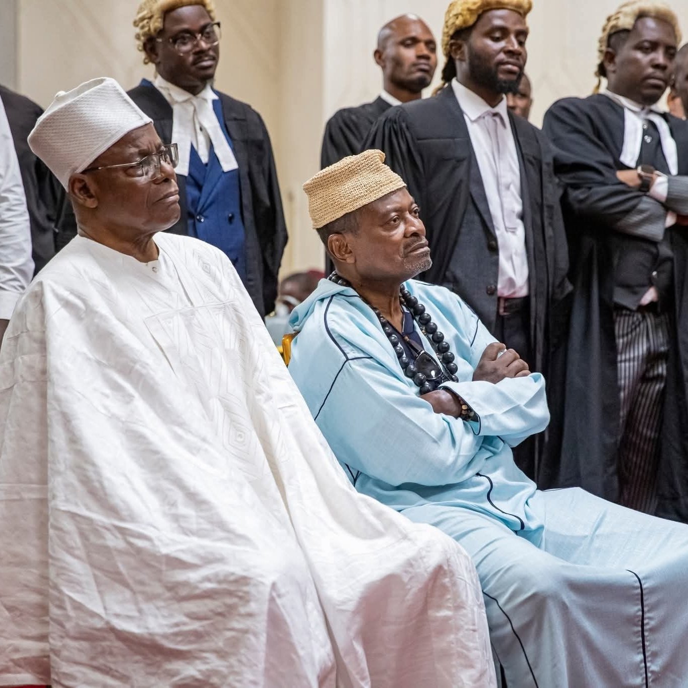 L'image montre un groupe de personnes rassemblées dans un espace formel, probablement un tribunal ou un événement solennel. Au premier plan, deux hommes sont assis. L'un porte une tenue traditionnelle blanche avec un chapeau, tandis que l'autre est habillé d'une tunique bleue et d'un collier de perles. Autour d'eux se trouvent des hommes en costume noir, certains portant des robes d'avocat. Tous semblent attentifs et sérieux, ce qui suggère que l'événement en cours est d'une grande importance. L'ambiance générale peut être perçue comme solennelle, avec une concentration visible sur les visages des participants.
