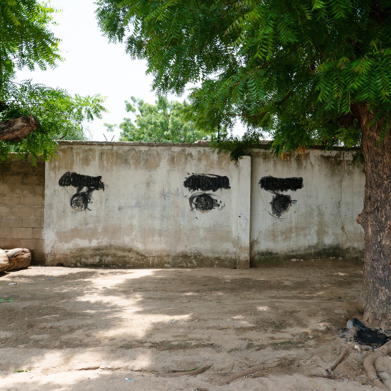 L'image présente un mur en béton aux couleurs neutres, probablement gris ou beige, partiellement recouvert par des ombres de feuillage provenant d'un grand arbre à proximité. Sur le mur, on peut voir des traces de peinture noire qui forment des motifs stylisés évoquant des yeux. Ces motifs sont positionnés de manière à créer une disposition particulière sur le mur. Au premier plan, on remarque également un tronc d'arbre robuste, avec des racines visibles qui émergent du sol sablonneux. L'environnement semble tranquille, avec une légère luminosité qui indique une journée ensoleillée.
