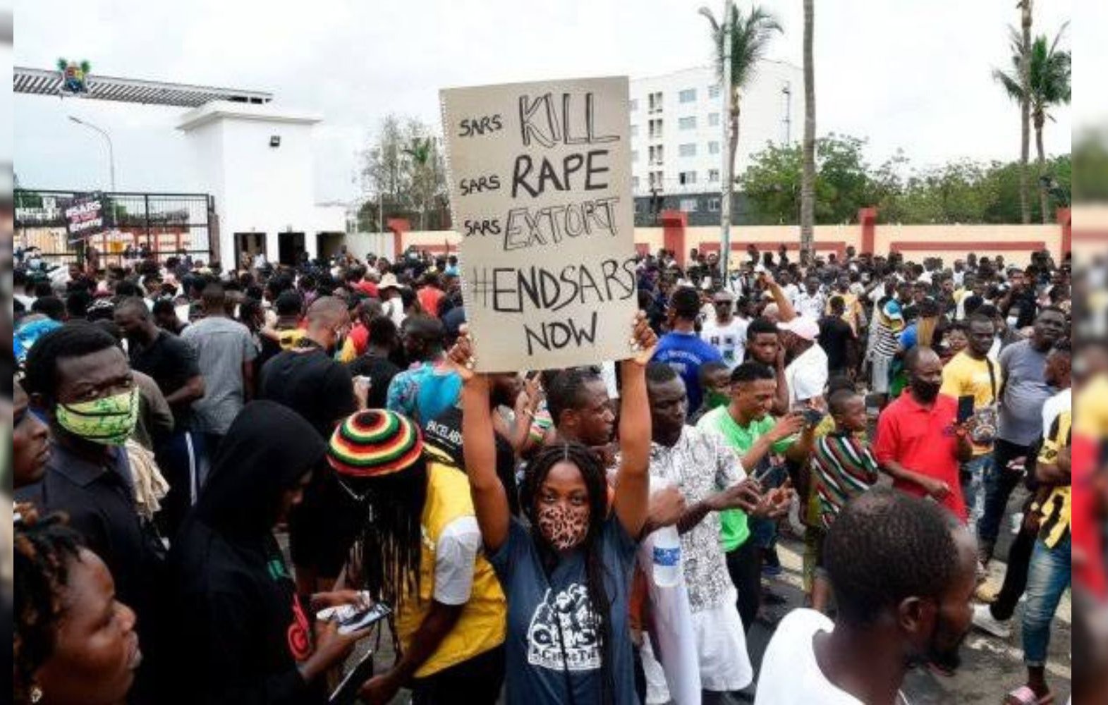 L'image montre une foule dense rassemblée pour une manifestation. Au centre, une personne tient une grande pancarte avec des inscriptions demandant la fin d'abus de la police. Les manifestants portent des vêtements variés, certains affichent des signes de solidarité, comme des masques. En arrière-plan, on aperçoit des bâtiments et des palmiers, ce qui donne une idée que l'événement se déroule dans une ville. L'atmosphère est chargée d'énergie collective, marquée par un sentiment de lutte et de revendication pour des droits.