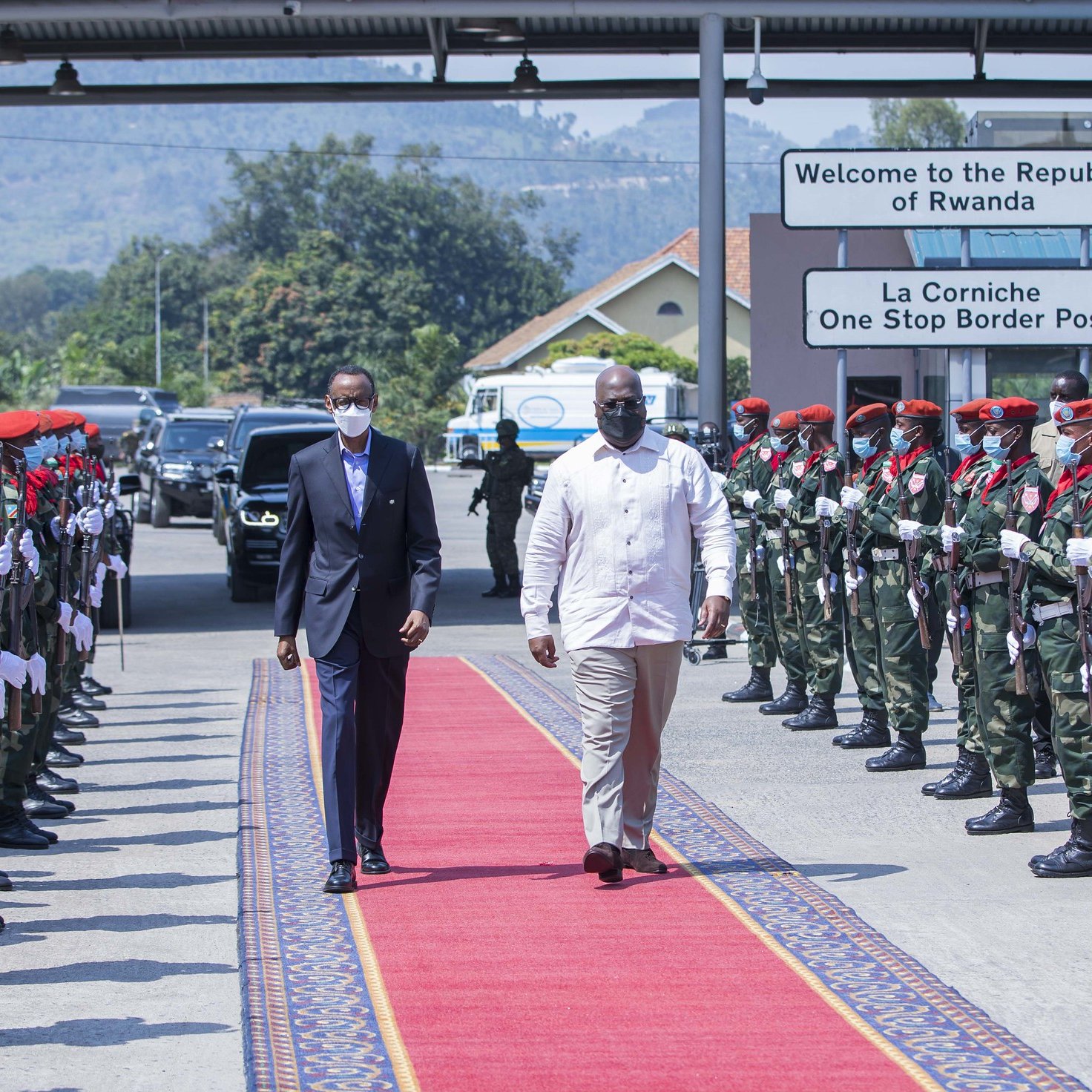 L'image montre un moment protocolaire à un poste frontalier. Deux hommes marchent sur un tapis rouge. À gauche, un homme en costume noir et masque, et à droite, un homme en chemise blanche. De chaque côté du tapis, des soldats en uniforme militaire attendent, formant une haie d'honneur. Les soldats portent des bérets rouges et des masques. En arrière-plan, on peut voir des panneaux indiquant la bienvenue au Rwanda et un poste frontière. L'ambiance est solennelle et cérémonielle.