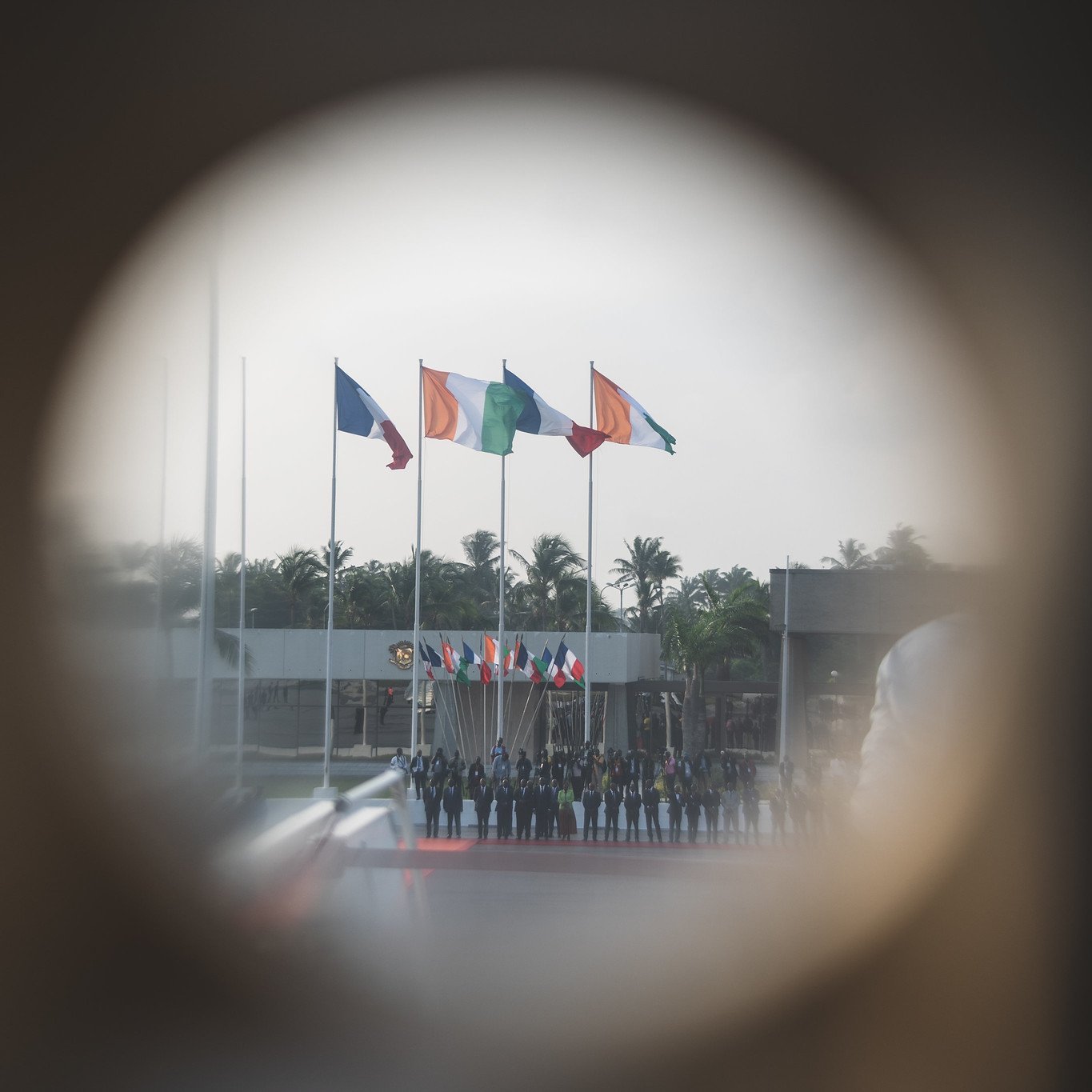 L'image montre un groupe de personnes alignées, probablement en uniforme, au centre d'un événement officiel. En arrière-plan, plusieurs drapeaux flottent au vent, représentant probablement différents pays. On distingue une ambiance solennelle, renforcée par la présence d'arbres aux feuilles verdoyantes et d'un espace dégagé. L'image est prise à travers un trou de serrure, ce qui donne une perspective unique et crée une atmosphère d'intimité et de mystère. La lumière semble douce et diffuse, suggérant un moment calme dans une journée ensoleillée.