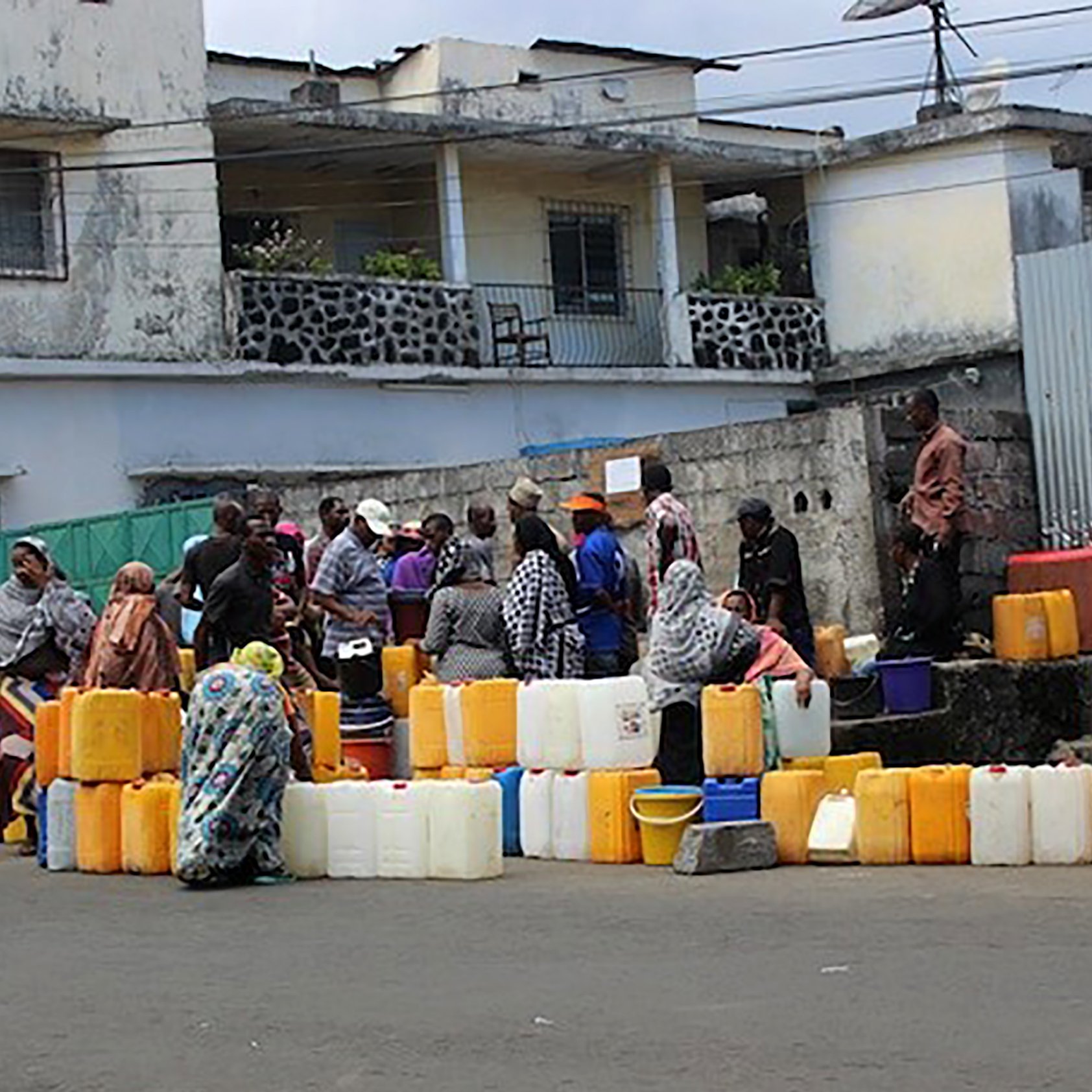 L'image montre une scène animée dans une rue. Des groupes de personnes se rassemblent autour de plusieurs conteneurs en plastique de différentes tailles, souvent de couleur orange ou jaune. Ces conteneurs sont alignés sur le sol. Les personnes, principalement habillées de vêtements traditionnels, semblent attendre leur tour pour remplir les récipients d'eau. En arrière-plan, on distingue des bâtiments aux façades simples, certains avec des balcons. L'ambiance est celle d'une activité quotidienne, soulignant l'importance de l'eau dans la vie des gens.