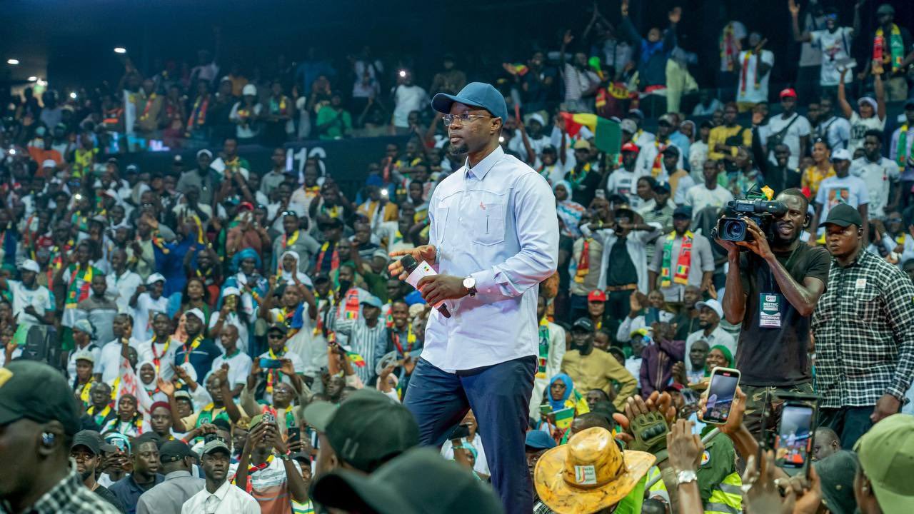 L'image représente un événement animé dans une grande salle remplie de personnes. Au premier plan, un homme en tenue décontractée, portant une chemise claire et une casquette, marche avec assurance sur la scène. Il semble interagir avec le public, qui se compose de nombreux spectateurs enthousiastes, certains levant les mains en signe d'encouragement. La foule est colorée, avec des personnes habillées en diverses tenues, et des drapeaux ou accessoires visibles, suggérant un fort esprit communautaire. En arrière-plan, on peut voir des caméras et des membres de l'équipe de sécurité, ce qui indique que l'événement est de grande envergure. L'atmosphère est d'excitation et d'engagement collectif.