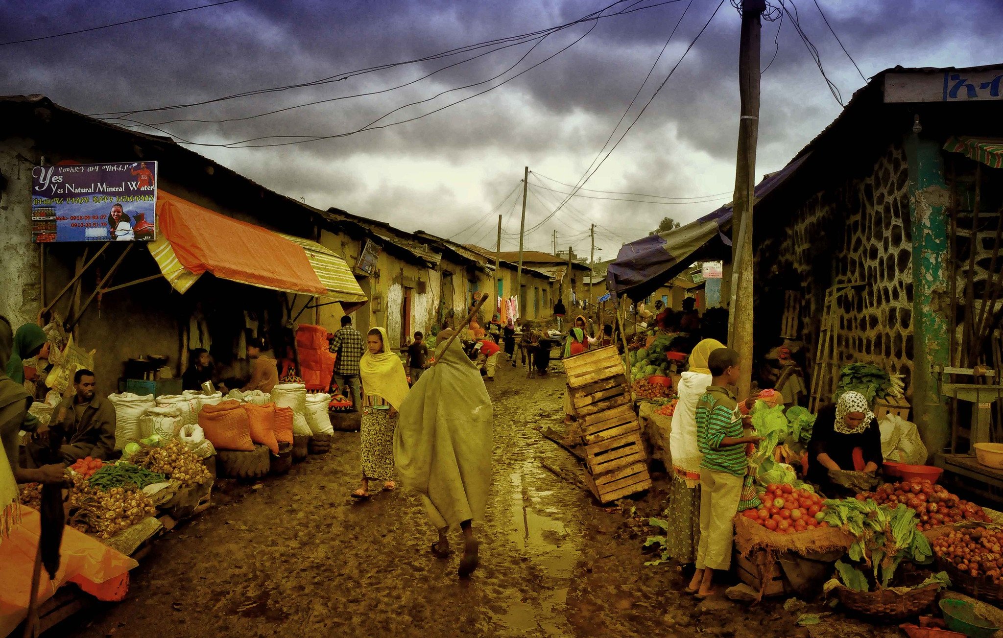 Cette image dépeint une scène de marché dans une rue étroite et animée. Le sol est boueux, probablement à cause de la pluie récente. Des étals colorés, couverts de bâches orange et bleues, sont remplis de fruits et légumes frais. Des vendeurs, portant des vêtements traditionnels et des capuchons pour se protéger de l'humidité, interagissent avec les clients. On peut ressentir l'effervescence du marché, avec des gens qui marchent, négocient et échangent des sourires. Au fond, le ciel est nuageux, ajoutant une atmosphère grise à cette scène vivante. Ce mélange de sons, d'odeurs et d'activités crée une ambiance dynamique et chaleureuse, typique des marchés locaux.