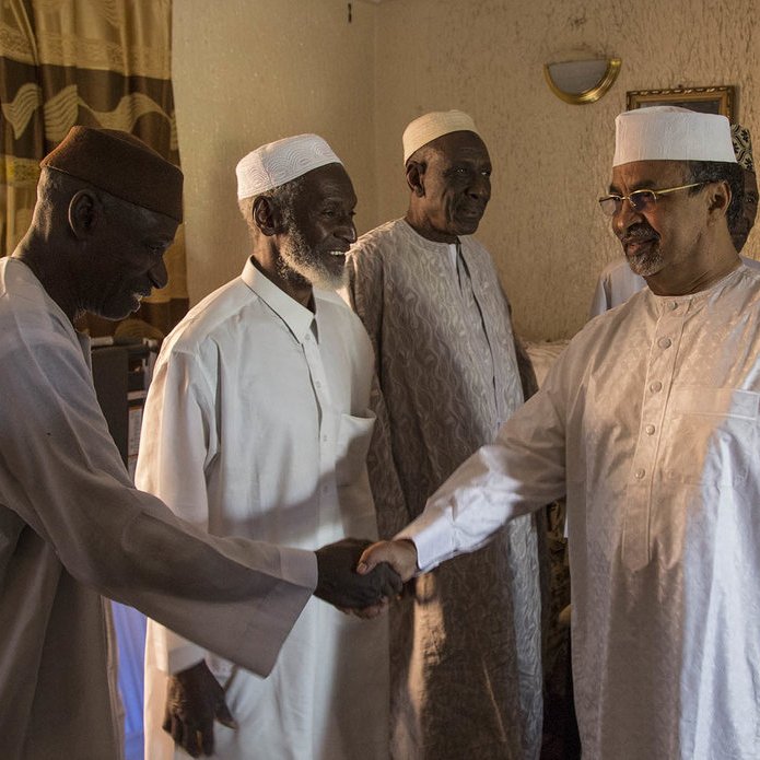 L'image montre un groupe de quatre hommes dans une pièce intérieure. Ils portent des vêtements traditionnels : des turbans et des vêtements clairs, souvent en coton. Un des hommes, sur la droite, est en train de serrer la main d'un autre homme, tandis que deux autres les regardent avec un air amical. La lumière entre par une fenêtre derrière eux, créant une atmosphère chaleureuse. Les murs sont texturés, avec un éclairage doux, et l'ensemble donne une impression de convivialité et de respect mutuel.