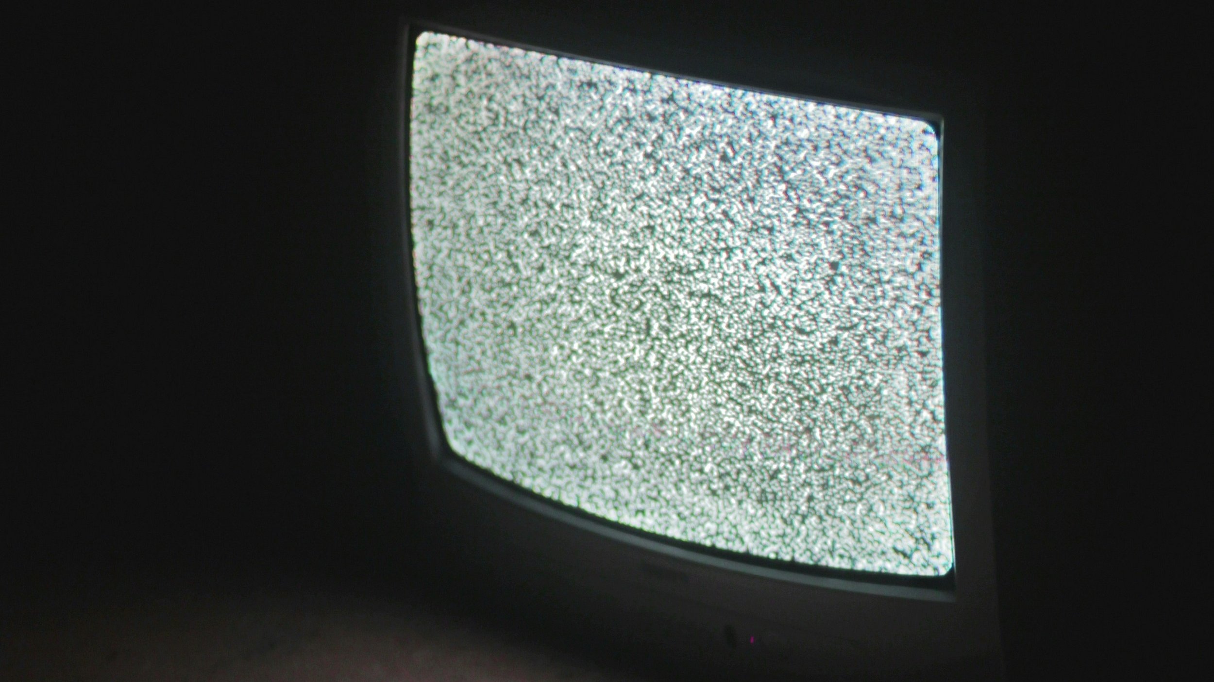 L'image présente un téléviseur ancien, dont l'écran affiche un bruit visuel, souvent appelé "neige", constitué de points blancs et noirs qui scintillent. L'appareil est placé sur le sol, et l'éclairage ambiant est faible, ce qui met en avant le contraste de l'écran lumineux par rapport à l'obscurité environnante. Le téléviseur a des bords arrondis et un design rétro, typique des modèles d'une autre époque. La scène dégage une atmosphère nostalgique et un sentiment d'attente.