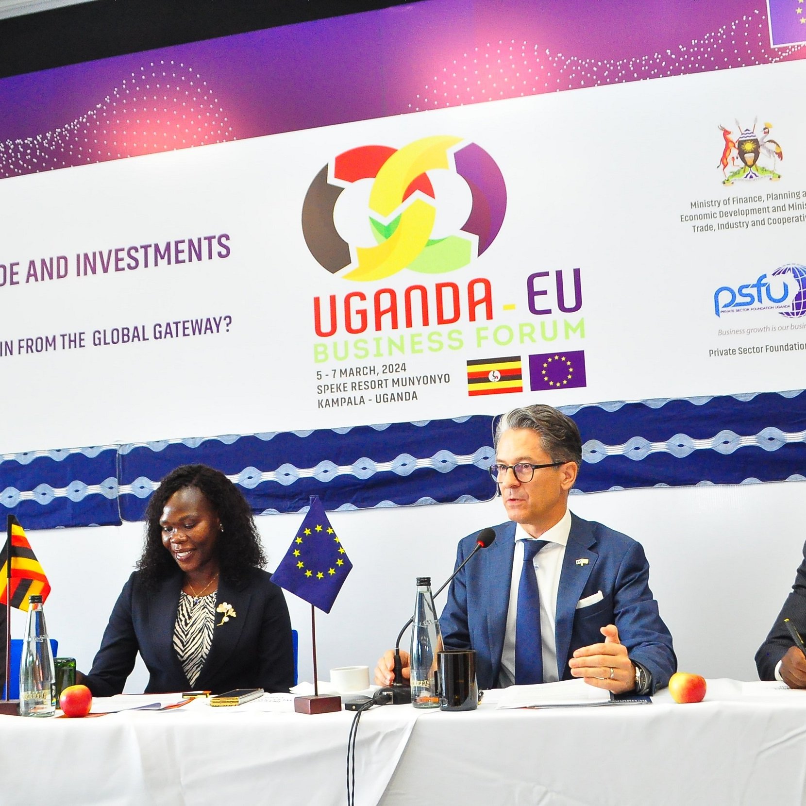 L'image montre une scène d'un forum d'affaires intitulé "Uganda-EU Business Forum". Au premier plan, quatre personnes sont assises à une table, chacune ayant un micro devant elle. À gauche, un homme souriant semble représenter l'Ouganda. Ensuite, une femme, également souriante, prend la parole. Au centre, un homme, avec des lunettes et un air sérieux, est en train de s'exprimer. Enfin, à droite, un autre homme, avec une posture attentive, écoute. En arrière-plan, une banderole avec des logos et le titre de l'événement est accrochée. L'ambiance est professionnelle, et l'événement semble axé sur le renforcement des échanges et des investissements entre l'Uganda et l'Union européenne.