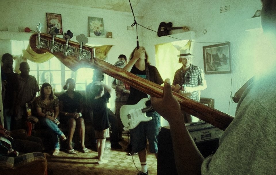 L'image montre une scène musicale vivante dans une pièce chaleureuse. À l'avant, on voit un musicien tenant une basse, tandis qu'un autre, avec une guitare électrique, se concentre sur sa performance. Les membres du groupe sont entourés d'un public qui semble apprécier la musique. Les visages sont tournés vers les musiciens, certains se balançant au rythme. La pièce est décorée avec des tableaux aux murs et des rideaux qui laissent filtrer une lumière douce. L'ambiance est conviviale, remplie d'énergie et de passion pour la musique.