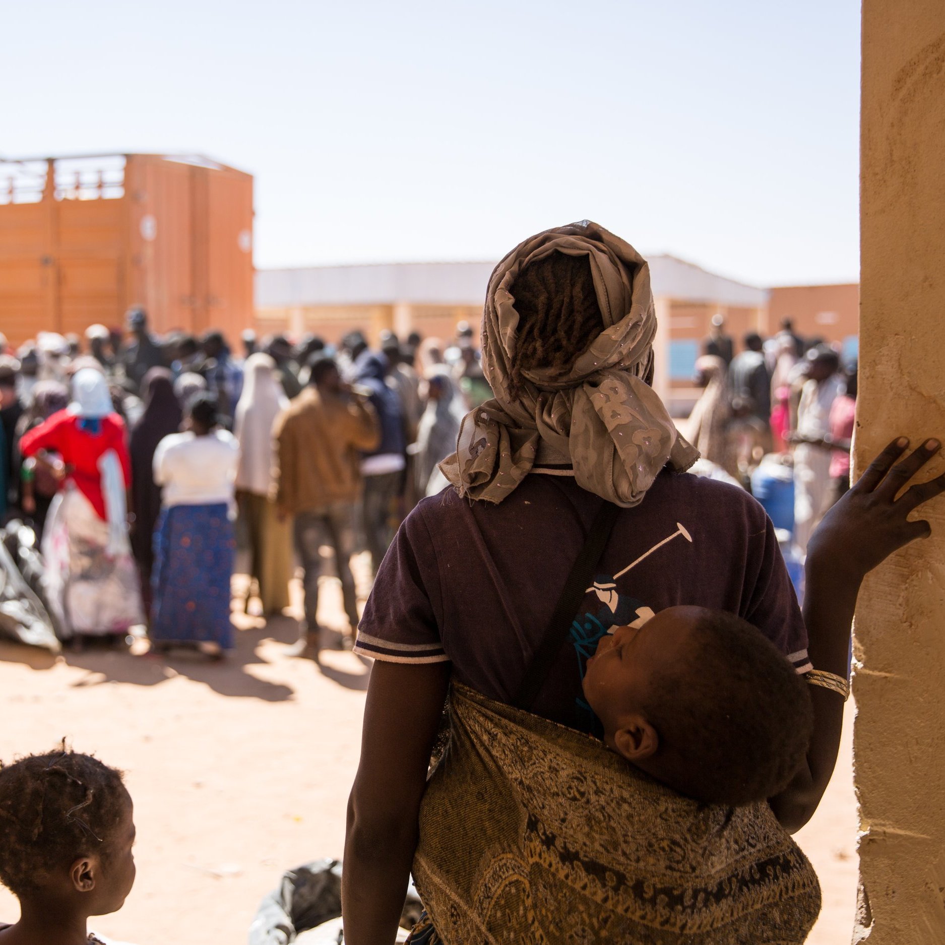 L'image montre une scène poignante dans un contexte de rassemblement de personnes, probablement dans un camp ou un centre d'accueil. Au premier plan, une personne, probablement une mère, porte un enfant dans un sling. Elle regarde vers un groupe de personnes, qui attendent patiemment, formant une masse compact de silhouettes. La majorité des personnes dans le fond sont habillées de vêtements aux couleurs variées, certaines portant des foulards ou des vêtements traditionnels. Le sol est en terre battue, et des containers peuvent être aperçus à l'arrière-plan, suggérant un environnement temporaire. L'atmosphère semble à la fois calme et chargée d'une certaine tension, reflétant une réalité complexe.