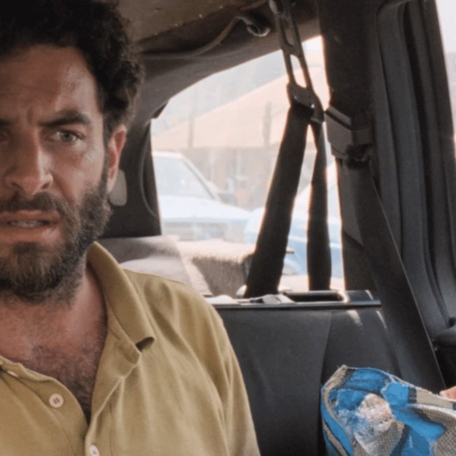 The image shows two people inside a vehicle. One person, likely wearing a blonde wig and sunglasses, leans towards the other, who has a beard and a concerned expression. The setting appears casual and slightly cramped, with a background that suggests they are parked in a busy area. The overall mood seems tense or serious, as both individuals have focused looks on their faces.