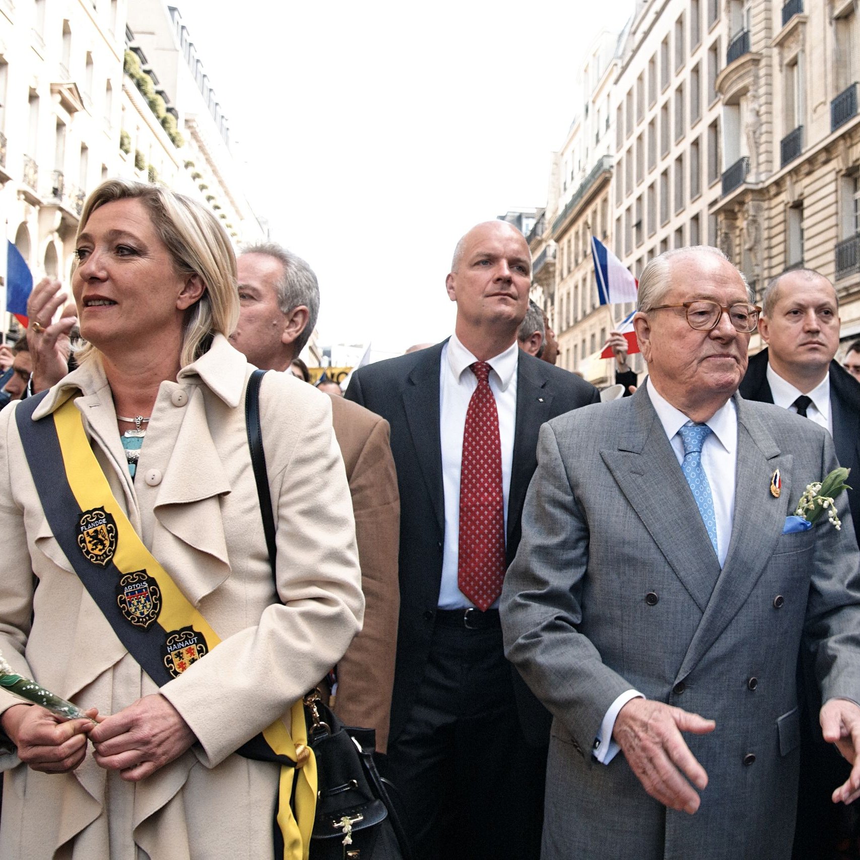 L'image montre un groupe de personnes marchant dans une rue animée, probablement lors d'un événement ou d'une cérémonie. Au centre, un homme plus âgé porte un costume gris clair et des lunettes. Il semble être une figure de proue, entouré de plusieurs autres individus, dont certains portent des sashes ou des écharpes distinctives, suggérant qu'ils occupent des rôles importants. On remarque également des décorations florales, indiquant une atmosphère cérémonielle. En arrière-plan, on aperçoit des bâtiments typiques, et des drapeaux flottent, ce qui évoque un rassemblement public festif et patriotique.