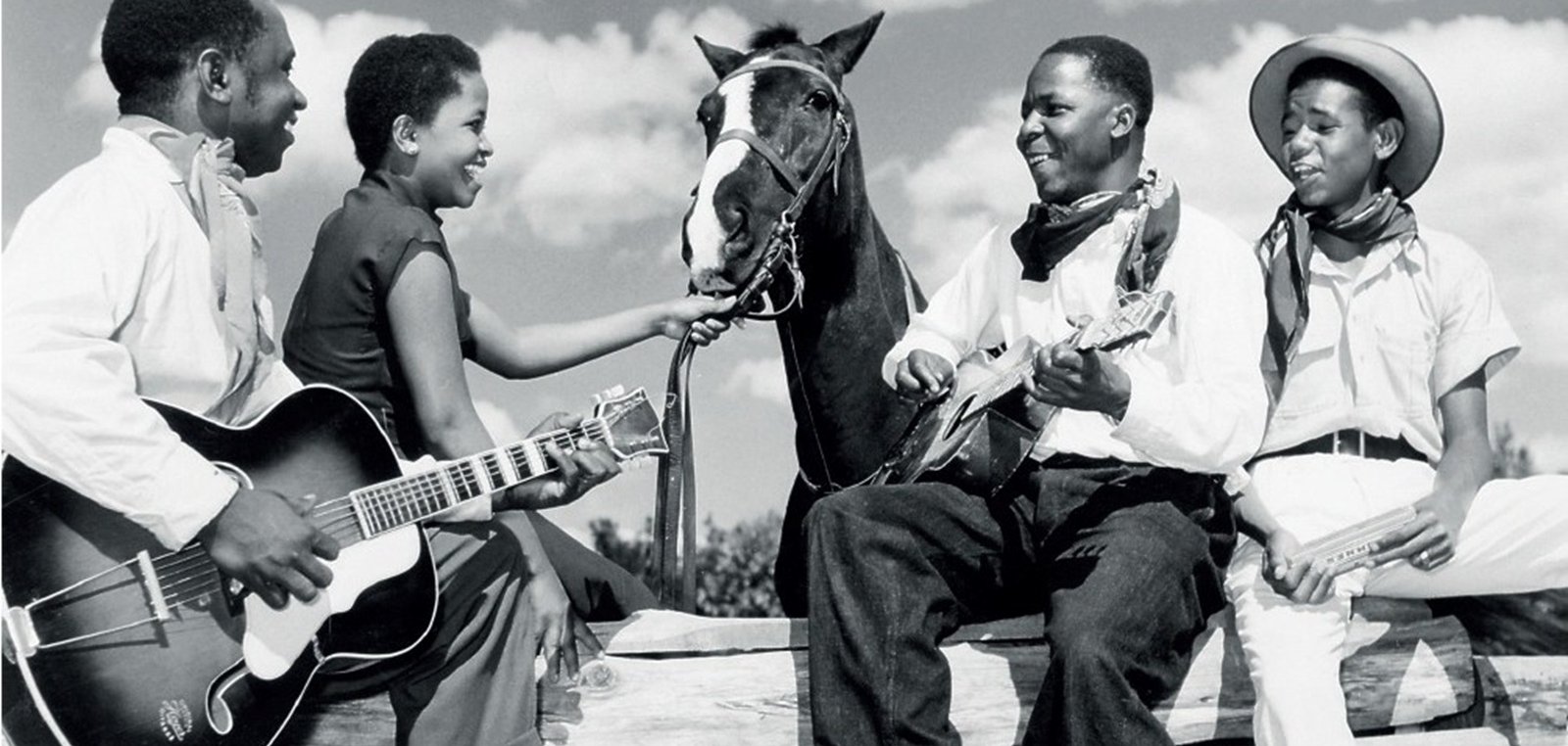 L'image représente un groupe de quatre personnes en noir et blanc, assises autour d'une barrière en bois, sous un ciel partiellement nuageux. Deux hommes et deux femmes sont présents. L'un des hommes joue de la guitare, tandis que l'autre est assis sur un tronc, tenant un instrument à cordes, probablement un banjo ou un ukulélé. Les deux femmes semblent interagir avec l'un des hommes et un cheval qui est également présent, la tête tournée vers eux. Elles affichent des expressions joyeuses, créant une atmosphère conviviale et détendue. Les vêtements des personnages varient : l'un porte un chapeau de cowboy et les autres portent des vêtements simples, typiques de l'époque. L'image évoque un moment de camaraderie et de musique, suggérant un lien fort entre les personnes et la nature environnante.