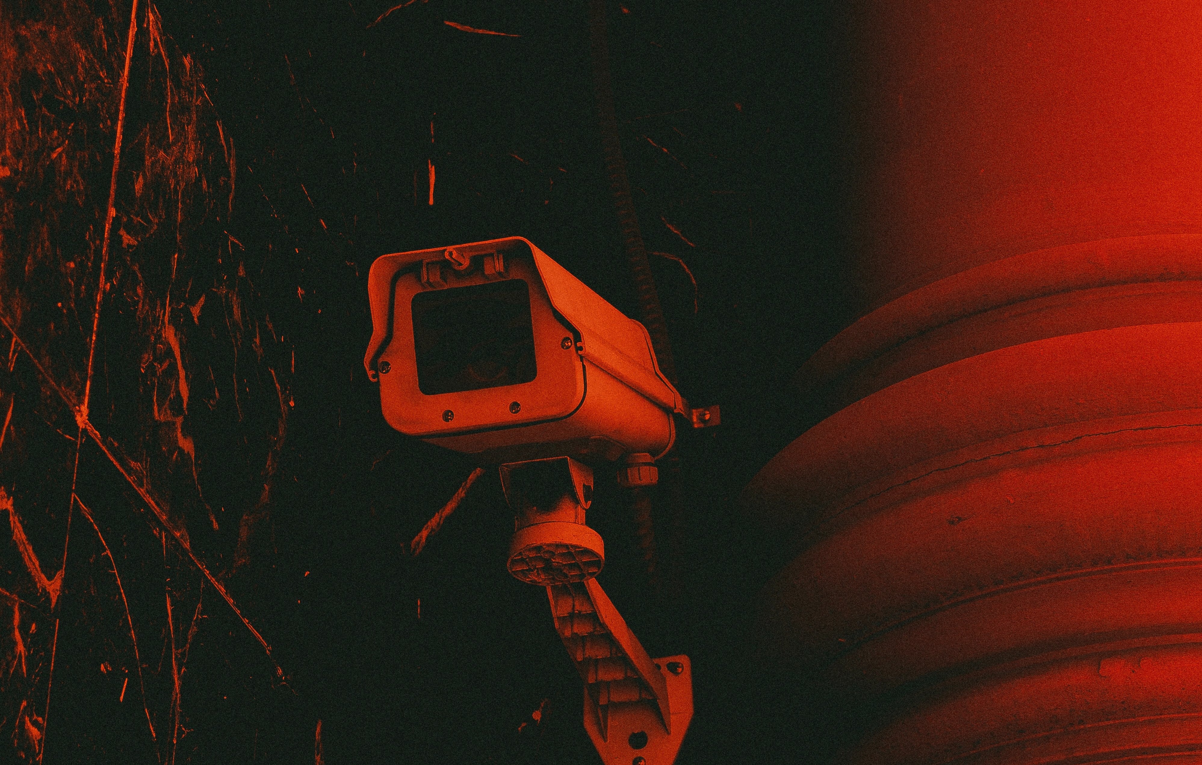 L'image représente une caméra de surveillance fixée à un mur. La caméra, avec son boîtier rectangulaire et son objectif, est peinte en gris clair. Elle est montée sur un support qui lui permet de pivoter, capturant ainsi une vaste zone de surveillance. L'arrière-plan est d'un rouge intense, créant une atmosphère sombre et légèrement inquiétante. Des textures sombres et des motifs abstraits se dessinent autour, ajoutant à l'effet dramatique de l'image. Cette combinaison de couleurs et de formes suggère une ambiance de vigilance et de sécurité.