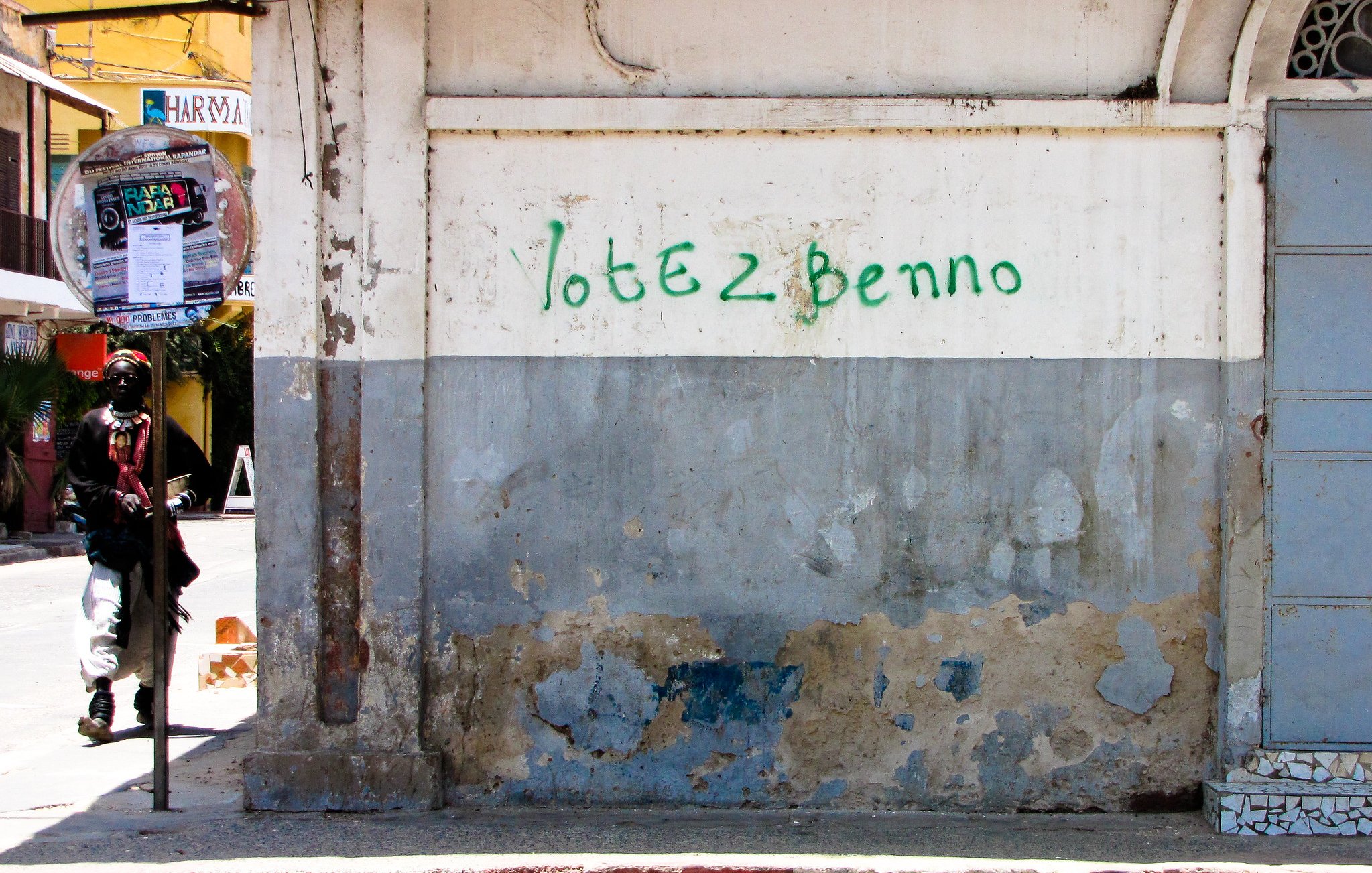 L'image montre un mur d'un bâtiment, principalement de couleur beige, avec des marques d'usure et de décoloration, témoignant de son ancienneté. Une inscription verte, "Votez Benno", est peinte sur le mur. À gauche, on aperçoit la silhouette d'un individu habillé de manière décontractée, qui semble marcher. Au fond, il y a une ambiance animée avec des éléments urbains tels que des affiches et des tables à l'extérieur de cafés. L'ensemble dégage une atmosphère de quartier vivant.