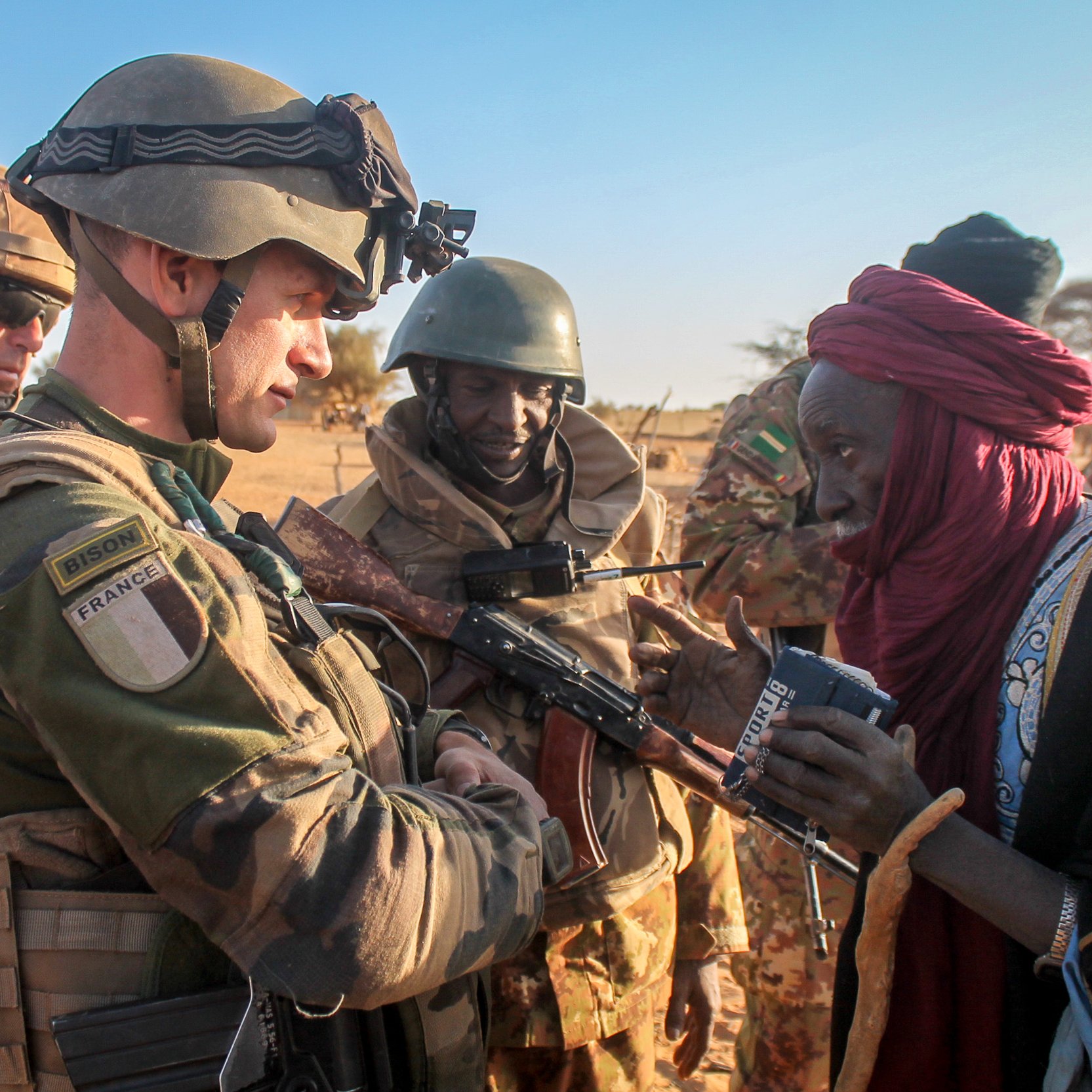 L'image montre une scène dans un environnement désertique, probablement au Sahel. Au premier plan, un soldat en uniforme militaire, portant un gilet pare-balles et un casque, interagit avec une femme vêtue d'une robe traditionnelle. Elle a l'air âgée et semble lui parler tout en tenant un objet dans ses mains. D'autres soldats en arrière-plan, arborant également des uniformes militaires, observent la scène avec un air attentif. L'éclairage est naturel, suggérant que la photo a été prise en fin d'après-midi, et le paysage environnant est sec avec des dunes de sable visibles. Cette rencontre reflète peut-être une interaction humanitaire ou de soutien entre les militaires et la population locale.