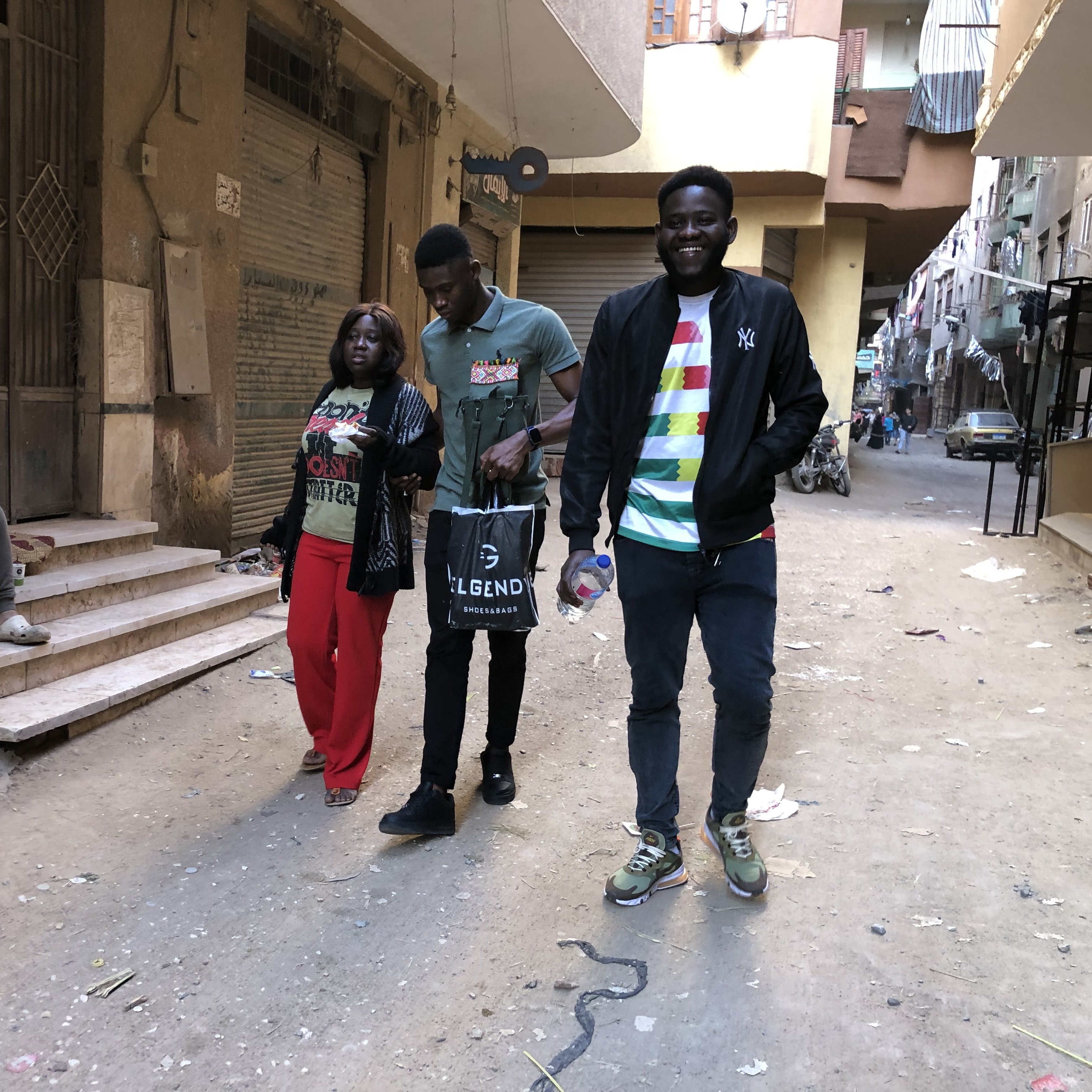 L'image montre une rue étroite et animée dans un quartier urbain. Il y a des déchets dispersés sur le sol, indiquant un environnement un peu désordonné. Sur le côté gauche, un jeune homme est assis sur des escaliers, absorbé par son téléphone ou ses réflexions. Au centre, trois personnes marchent ensemble : une femme portant un haut coloré et un pantalon rouge, un homme en t-shirt gris foncé et un autre homme vêtu d'un haut noir avec des détails colorés. Ils semblent converser joyeusement. En arrière-plan, on peut apercevoir plusieurs bâtiments qui témoignent de la vie locale, avec des balcons et des traces du quotidien. L'atmosphère est dynamique, capturant un moment de vie dans cette rue.