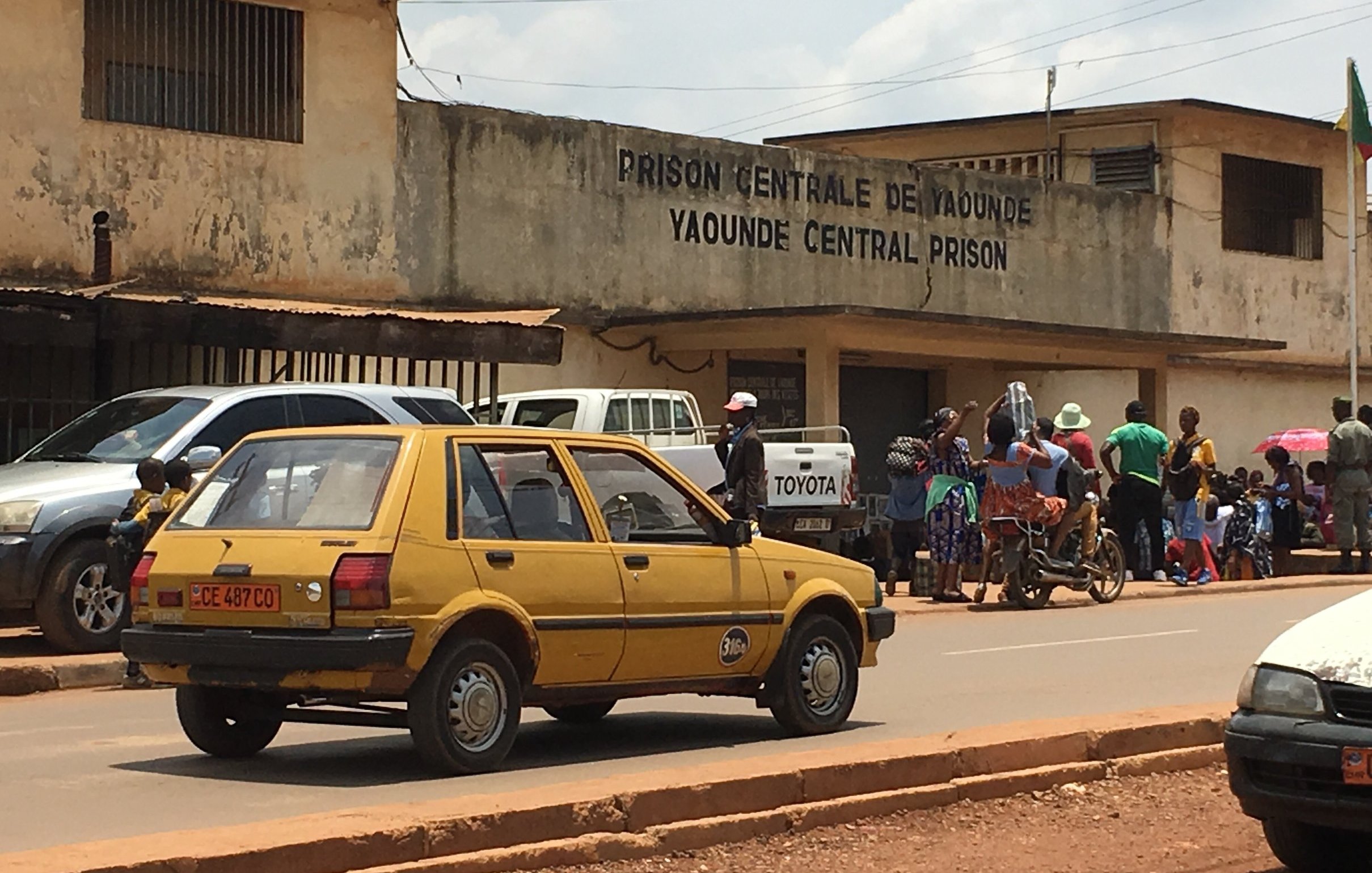 L'image montre l'extérieur de la prison centrale de Yaoundé, au Cameroun. On peut voir un bâtiment sobre avec des murs en béton, portant l'inscription "PRISON CENTRALE DE YAOUNDÉ" en lettres visibles sur la façade. En face de la prison, une route est occupée par une voiture jaune, typique des véhicules urbains. À côté, plusieurs personnes sont rassemblées, certaines discutant, d'autres attendant. Le décor autour est urbain, typique des villes africaines, avec une atmosphère animée. Le ciel est partiellement nuageux, ajoutant une nuance à la lumière de la scène.