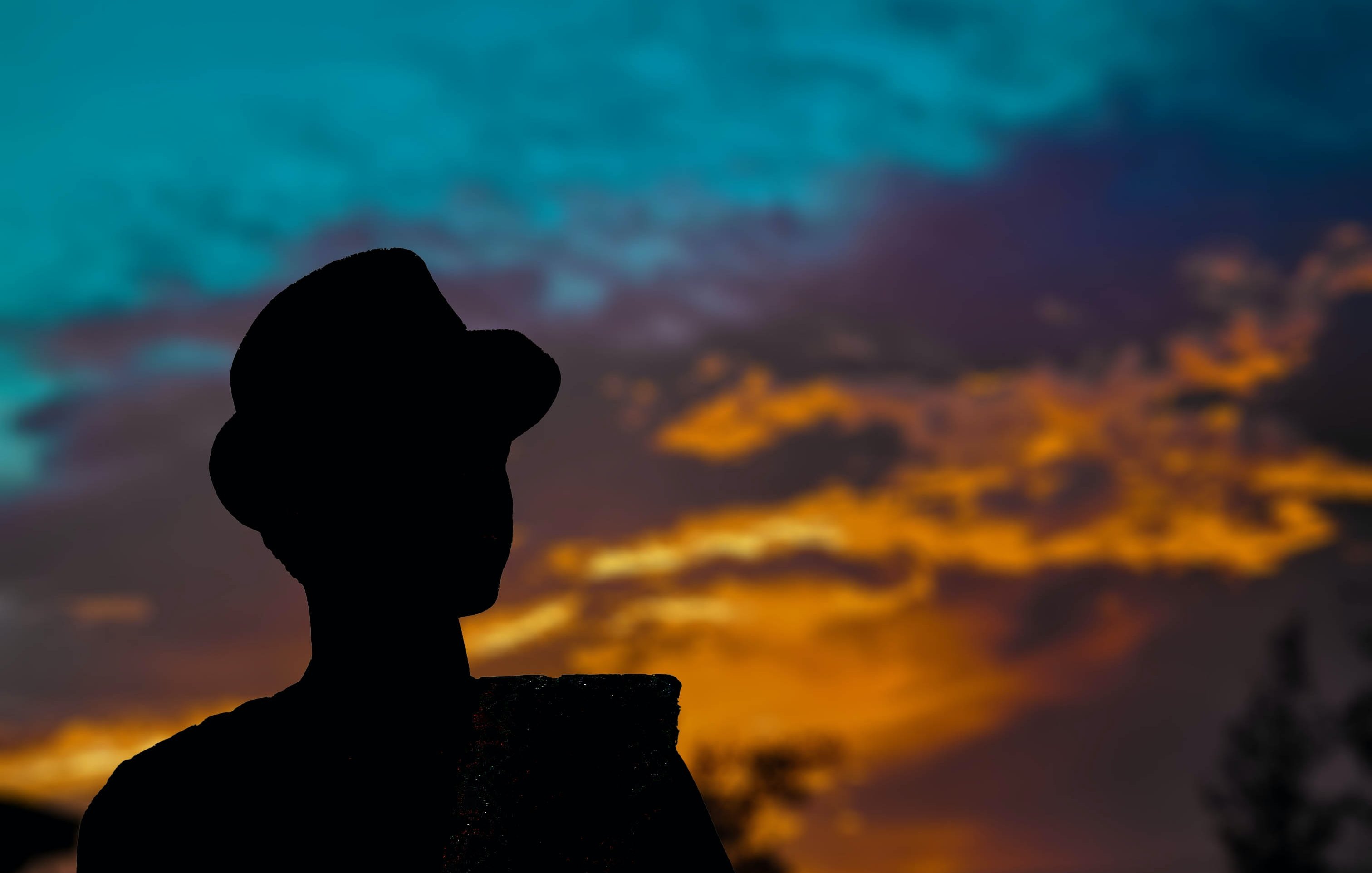 L'image présente une silhouette d'une personne se tenant debout, sur fond d'un ciel coloré au crépuscule. Le personnage porte un chapeau et semble contempler l'horizon. Le ciel est un mélange vibrant de bleu, d'orange et de violet, les nuages créant une atmosphère paisible et dramatique. Les couleurs chaudes du coucher de soleil se marient harmonieusement avec les teintes fraîches du ciel, engendrant une ambiance tranquille et contemplative.