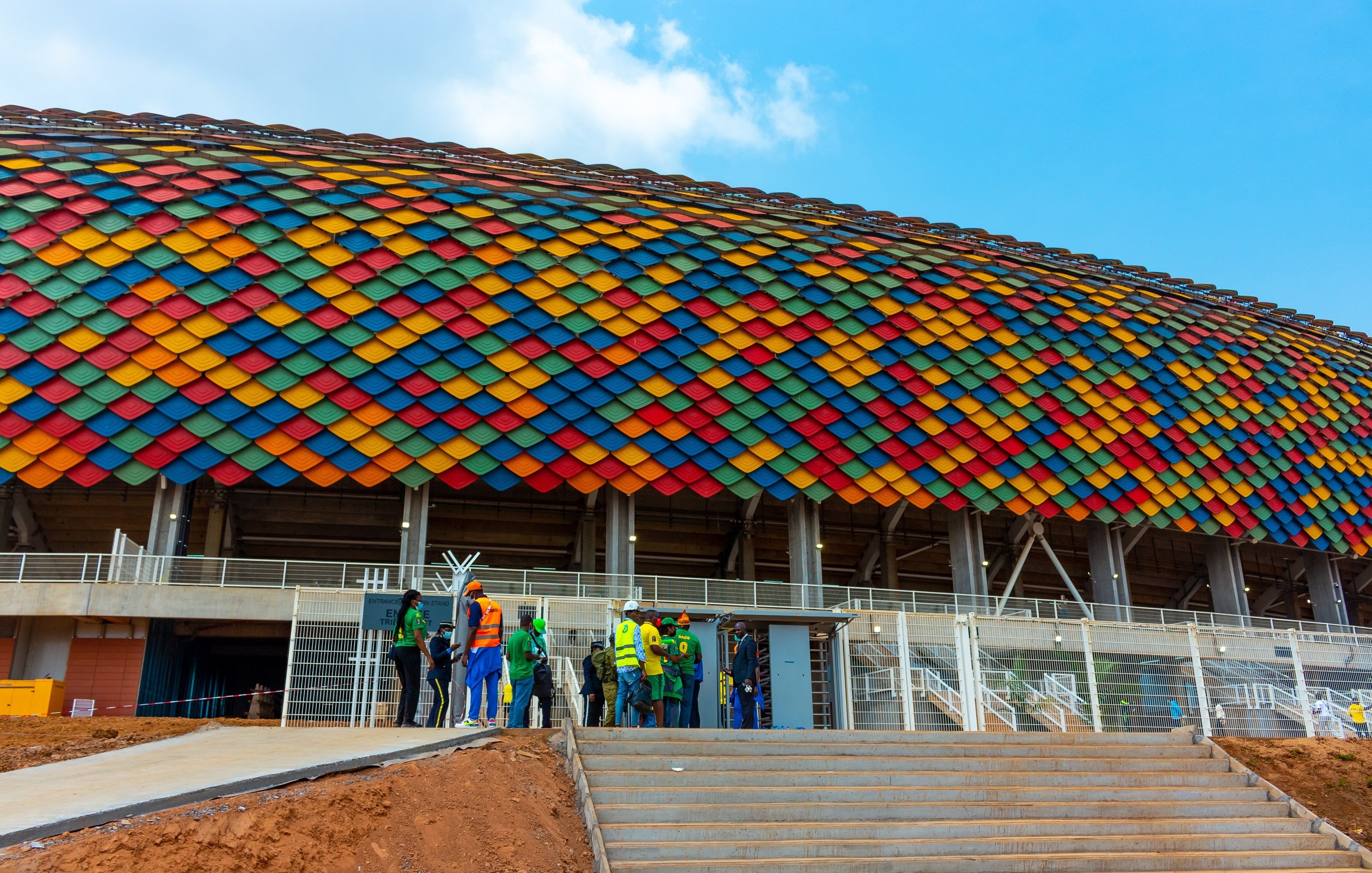 L'image montre un grand stade avec une architecture moderne. La façade du stade est ornée de tuiles de diverses couleurs vives, allant du rouge, orange, jaune, vert, bleu jusqu'au violet, formant une belle mosaïque sur un fond en béton. Au premier plan, on peut voir un groupe de personnes, habillées en vêtements de travail, qui semblent discuter ou se préparer à entrer dans le stade. Les escaliers mènent à une entrée principale, et le ciel au-dessus est bleu avec quelques nuages épars, ajoutant une impression de clarté et d'énergie à la scène.