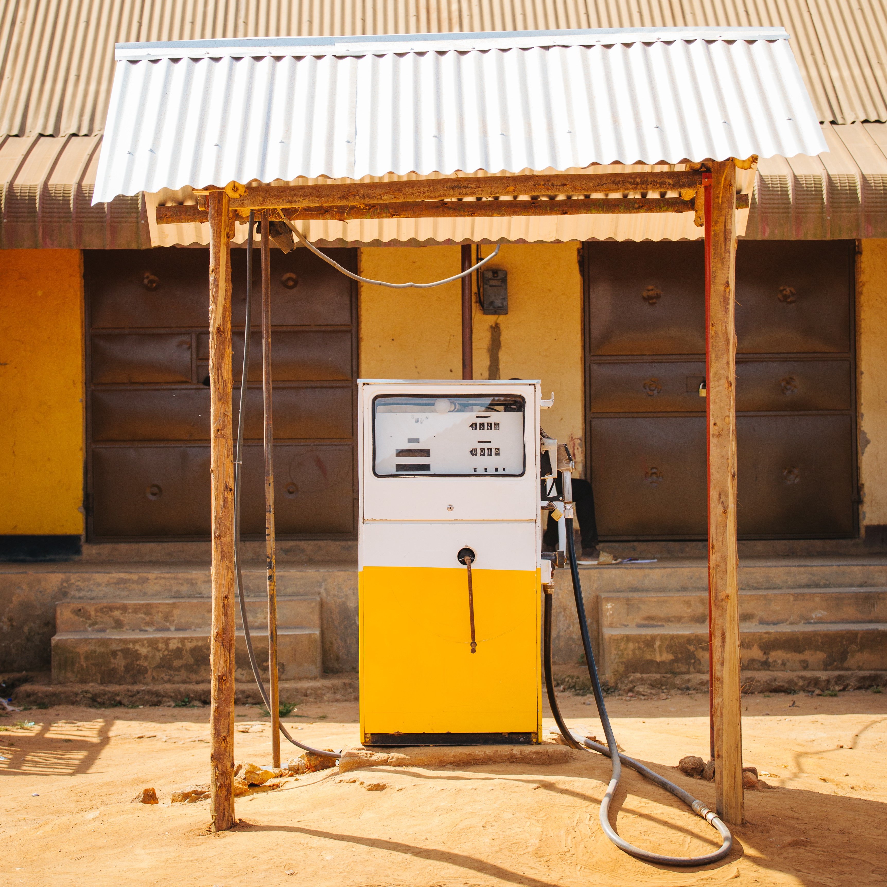Imaginez une petite station-service située à l'extérieur d'un bâtiment. Au centre de l'image se trouve une pompe à essence de couleur blanche et jaune, installée sous un auvent en tôle ondulée qui offre un peu d'ombre. La pompe est équipée d'un écran et d'un bouton pour le paiement, ainsi qu'un flexible noir reliant la pompe au réservoir d'un véhicule. À l'arrière-plan, on aperçoit un mur de couleur jaune avec deux portes en métal marron, fermées, et l'ensemble est posé sur un sol terreux, suggérant une ambiance rustique et simple. Le lieu semble tranquille, sans voitures visibles autour de la pompe.