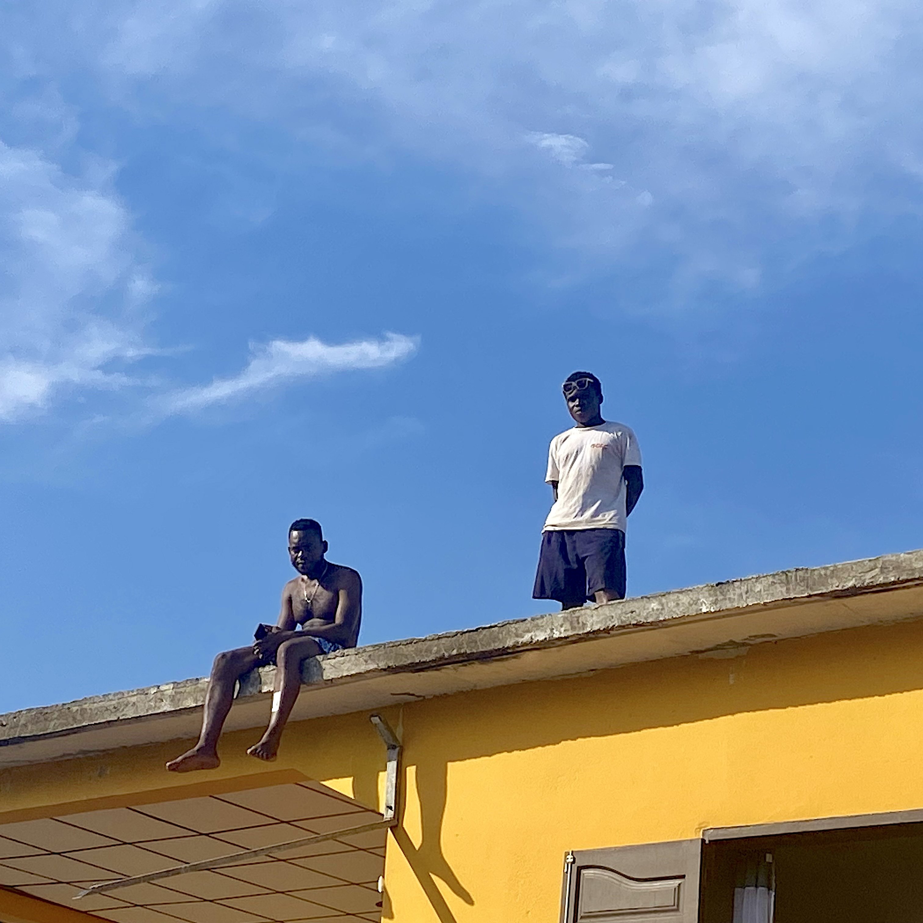 Sur cette image, deux jeunes hommes se trouvent sur le toit d'une maison peinte en jaune vif. L'un d'eux est assis au bord, les jambes pendantes dans le vide, tandis que l'autre se tient debout à ses côtés. Le ciel est clair et bleu, avec quelques nuages légers. L'ambiance semble détendue, peut-être un moment de loisirs ou de camaraderie. Les détails de l'environnement autour d'eux ne sont pas visibles dans cette capture, mais l'accent est mis sur leur posture et l'interaction entre eux.