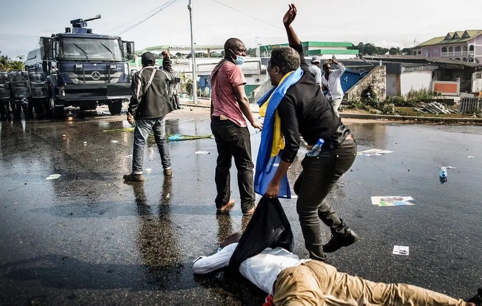 L'image montre une scène de manifestation dans une rue. Au premier plan, on peut voir des personnes en mouvement, certaines en train de gesticuler, tandis qu'une personne est à terre, apparemment blessée. L'un des manifestants porte un drapeau, probablement symbolique. En arrière-plan, il y a un véhicule de police, ce qui suggère une présence des forces de l'ordre. Le sol est couvert de débris, comme des affiches et des bouteilles, témoignant de la tension de la situation. L'ambiance semble intense et sérieuse, avec une impression de conflit.