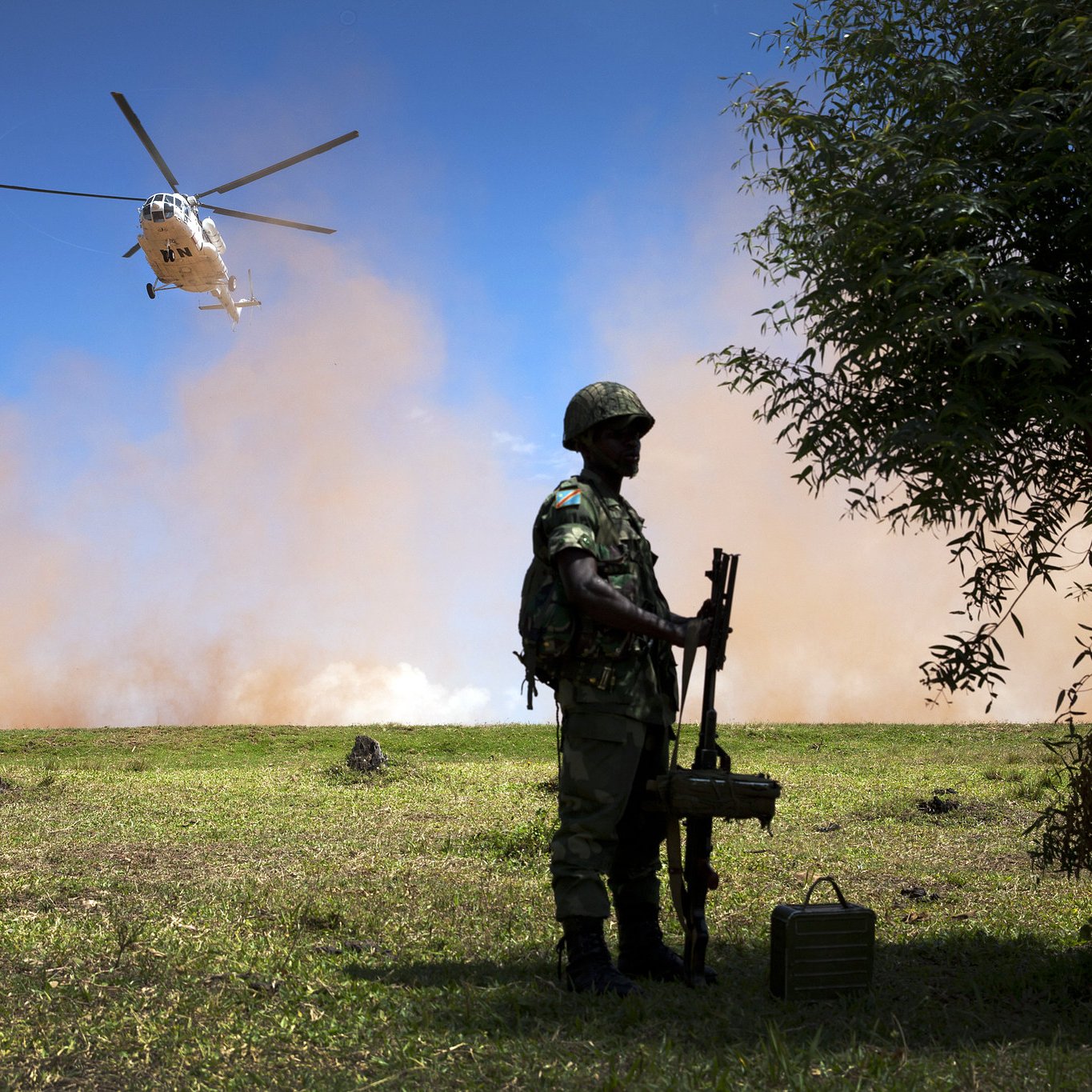 L'image représente un paysage militaire. Au premier plan, on voit un soldat debout, tenant une arme. Il porte un casque et un uniforme camouflage, suggérant qu'il se trouve dans un environnement opérationnel. À l'arrière-plan, un hélicoptère survole la scène, produisant un mouvement et une dynamique. Le ciel est clair, mais il y a également un nuage de poussière ou de fumée, ajoutant une atmosphère dramatique à la scène. Le terrain est verdoyant, mais des éléments comme des troncs d'arbres ou des obstacles peuvent être perçus, contribuant à l'ambiance militaire et à l'action en cours.