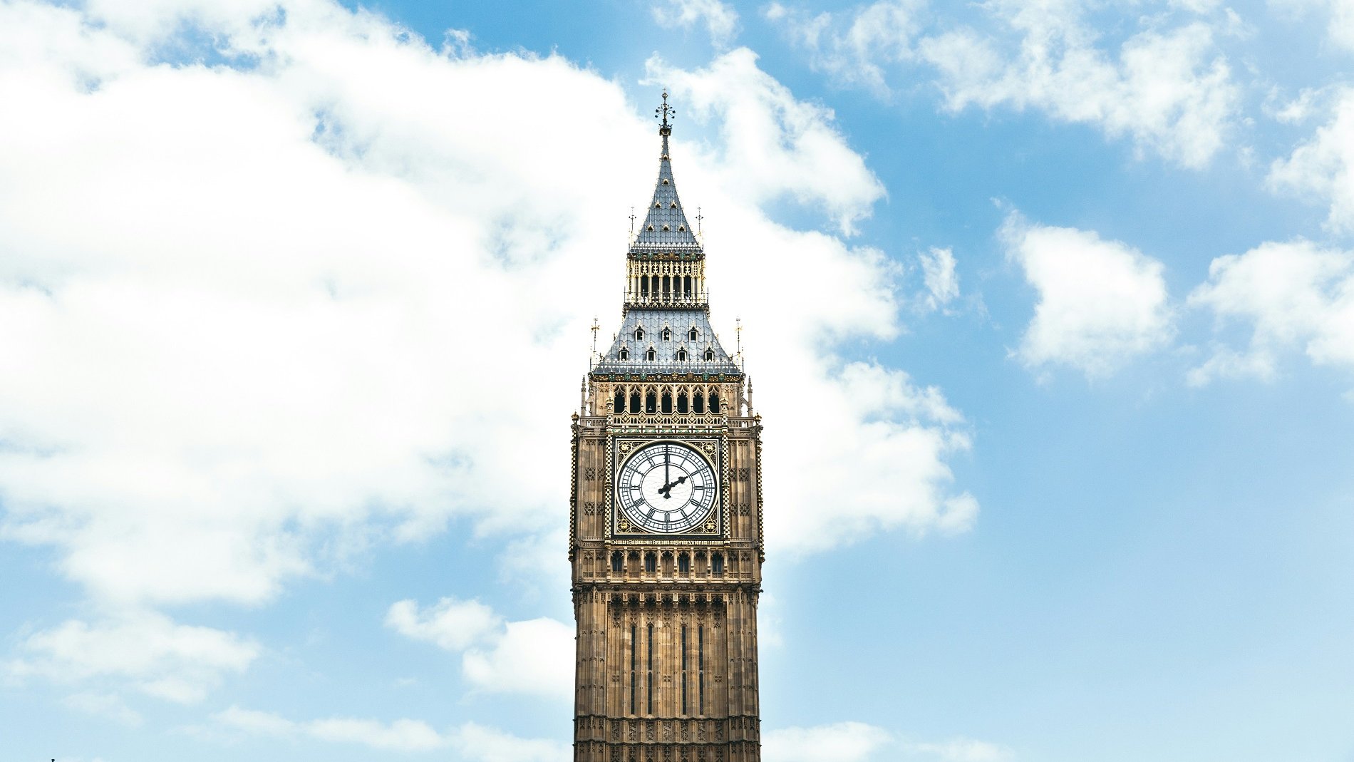 Imaginez un grand clocher en pierre, qui s'élève majestueusement vers le ciel. Ce bâtiment emblématique, connu sous le nom de Big Ben, est orné d'une grande horloge dont les aiguilles sont parfaitement visibles, indiquant l'heure avec précision. La tour est décorée de détails architecturaux, avec des voûtes et des motifs, et se dresse dans un ciel bleu parsemé de nuages blancs cotonneux. La lumière du jour accentue les couleurs chaudes de la pierre, ajoutant une touche rayonnante à cette structure historique.