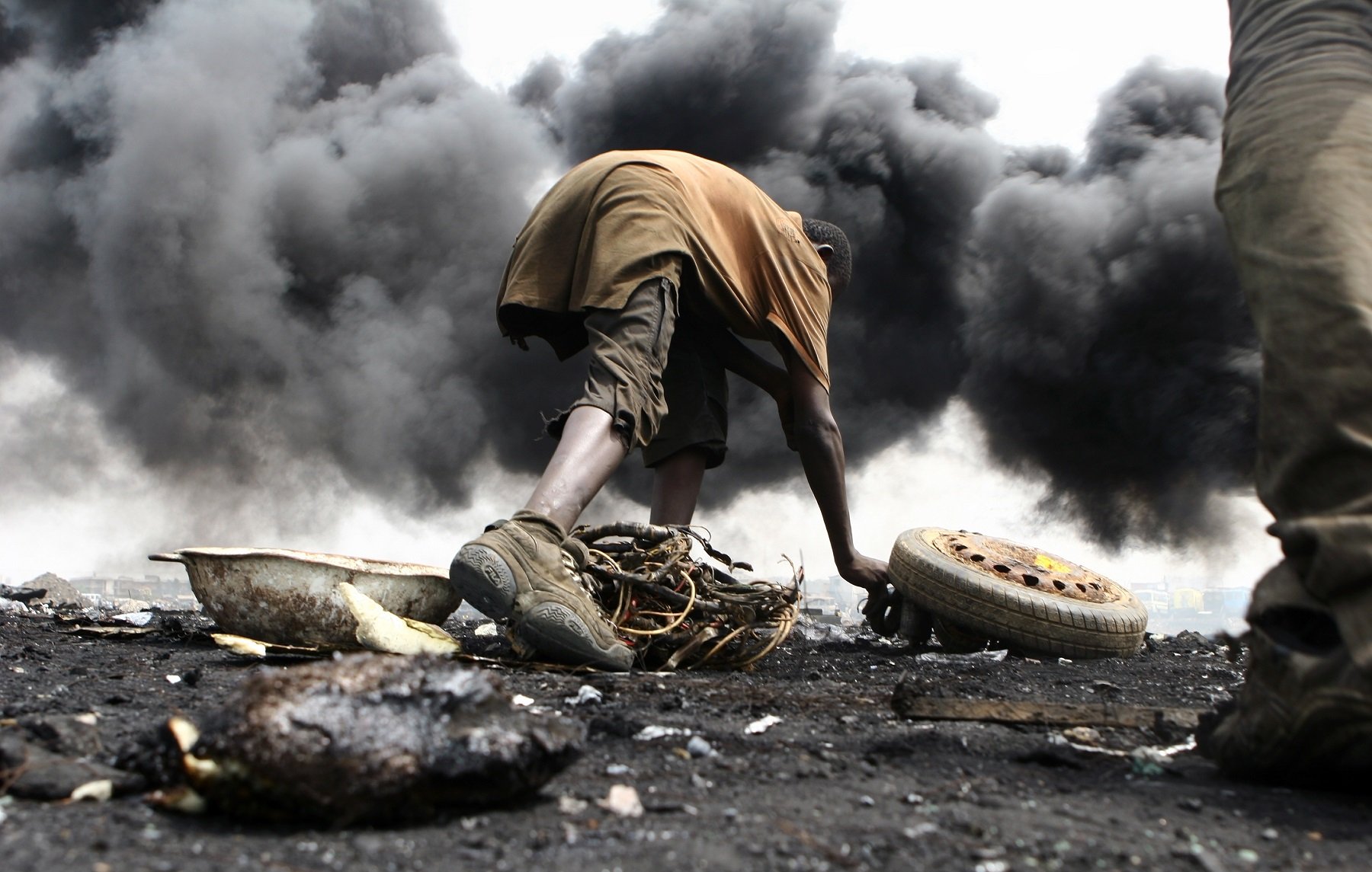 L'image montre un environnement très sombre et pollué, avec d'épaisses colonnes de fumée noire s'élevant dans l'air. Au premier plan, un jeune homme est accroupi, en train de manipuler des débris. Il porte des vêtements simples, à moitié sales, et semble concentré sur ce qu'il fait. Son environnement est chaotique, avec des morceaux de métal, de la ferraille et des pneus éparpillés autour de lui. La scène dégage une atmosphère de difficulté et de lutte, illustrant les conditions de vie dans cet endroit touché par la pollution et la dégradation environnementale.