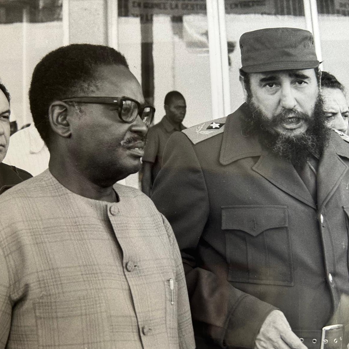 Cette image en noir et blanc montre trois hommes en discussion. Au premier plan, deux hommes sont particulièrement visibles. À droite, un homme avec une barbe fournie et un uniforme militaire, caractéristique de Fidel Castro, dialogue avec un homme portant des lunettes et un costume clair. Ce dernier semble attentif à la conversation. En arrière-plan, un troisième homme observe la scène. L'environnement est urbain, avec des bâtiments à vitre en arrière-plan. L'atmosphère paraît sérieuse et engagée, suggérant une rencontre diplomatique. Les expressions des visages laissent transparaître une certaine gravité.