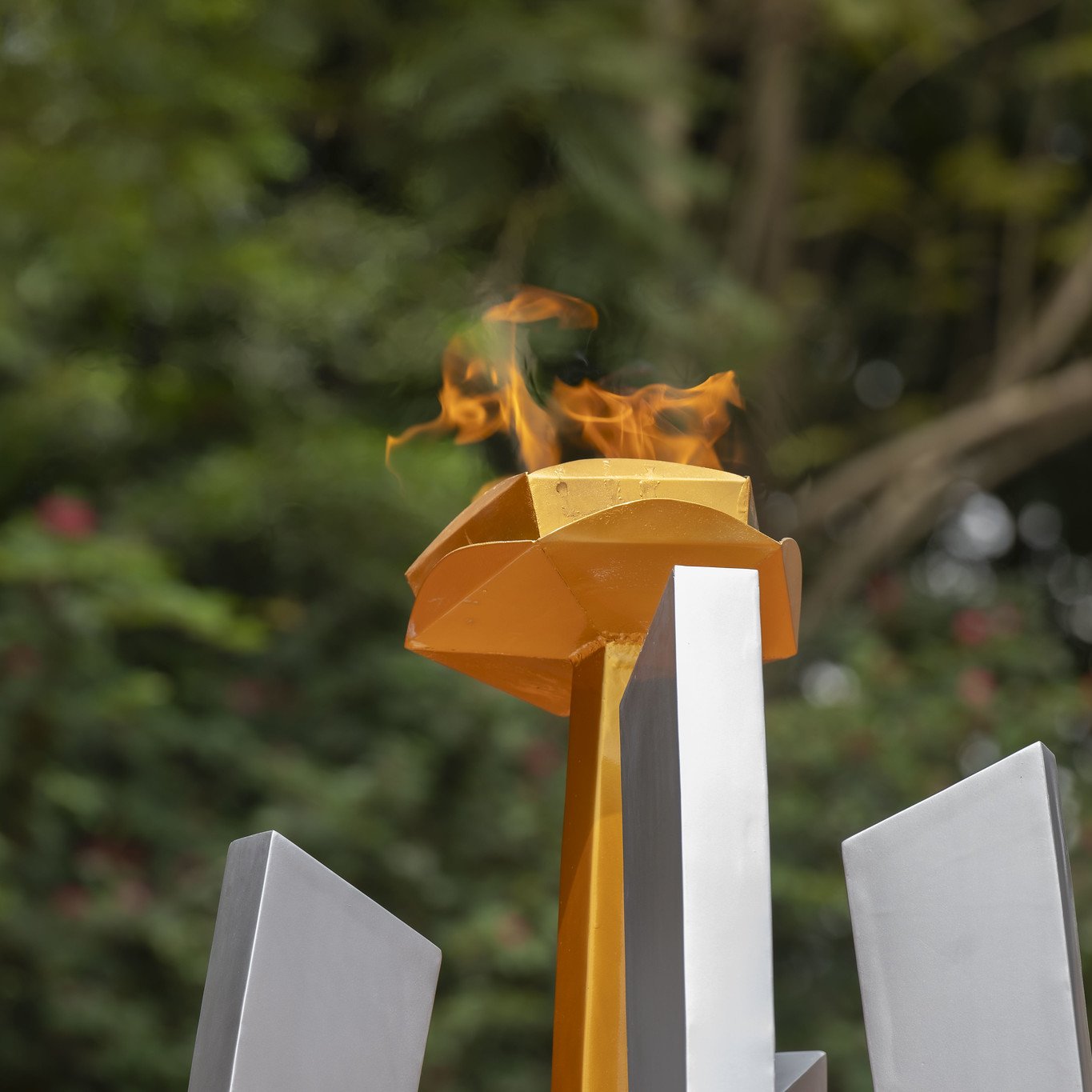 L'image représente une flamme allumée sur un socle jaune, qui surplombe plusieurs structures géométriques grises. La flamme danse avec vivacité, créant une ambiance dynamique, entourée d'un fond verdoyant flou, suggérant la présence d'arbres et de végétation. Cette scène évoque un symbole de passion et de célébration, souvent associé à des événements prestigieux comme les Jeux Olympiques. L'ensemble dégage une impression de force et de détermination.
