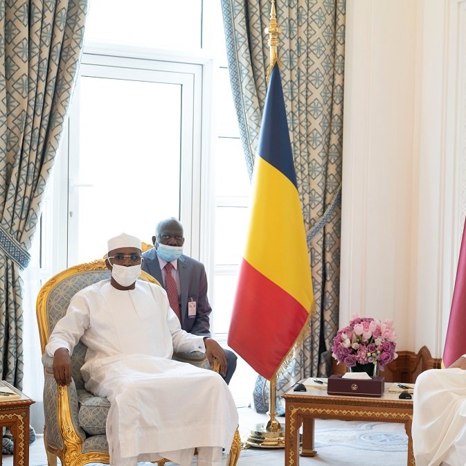 L'image montre une réunion formelle entre deux dirigeants. À gauche, un homme porte une tenue traditionnelle blanche et un chapeau, symbole de son pays. À sa droite, un autre homme est habillé d'une robe longue beige et d'un couvre-chef traditionnel. Entre eux, une table basse en bois contient des objets décoratifs. En arrière-plan, on peut voir des drapeaux : celui du Tchad à gauche et celui du Qatar à droite. La décoration des murs et des rideaux est élégante, avec des motifs riches et des couleurs apaisantes. L'atmosphère est solennelle et professionnelle.