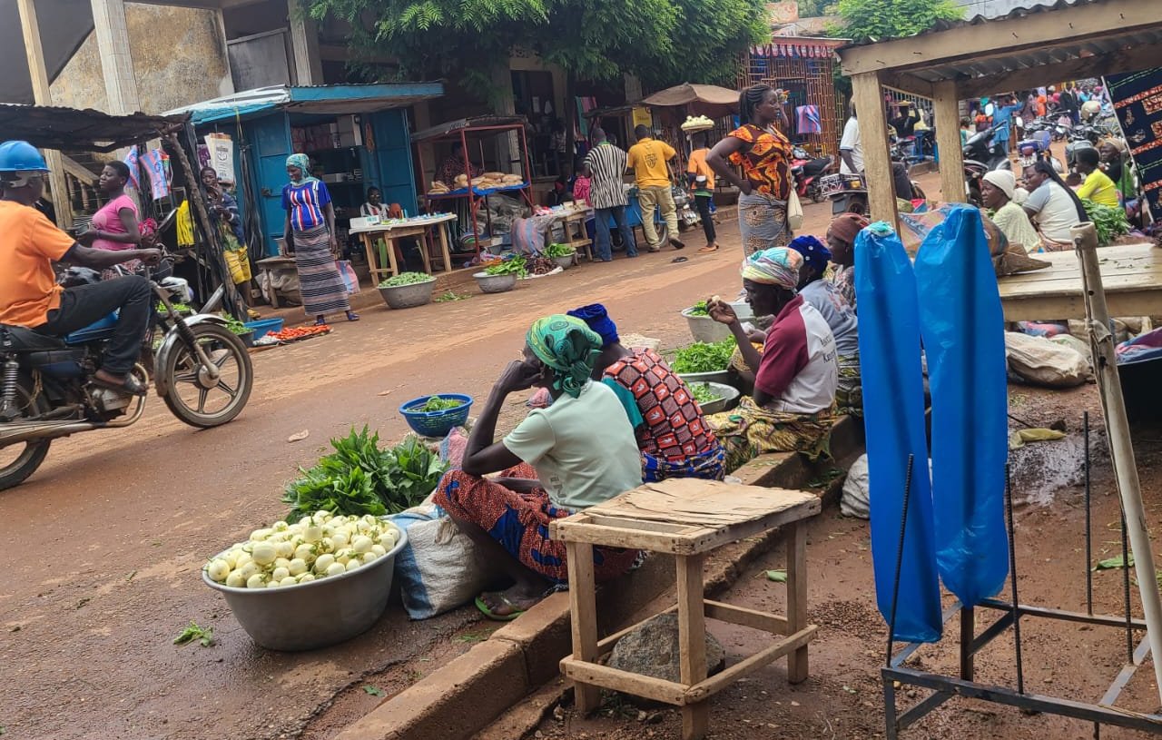 L'image montre une scène de marché vivant. Sur un chemin en terre battue, des femmes sont assises sur des banquettes en bois, entourées de paniers et de légumes frais. Certaines des femmes portent des tissus colorés et des foulards sur la tête, et elles semblent occupées à vendre des produits comme des légumes verts. En arrière-plan, des vendeurs s'affairent et des clients circulent. Des motos passent également sur la route, ajoutant à l'animation du lieu. L'atmosphère générale est vibrante, illustrant la vie quotidienne d'un marché local. Des arbres feuillus offrent une ombre bienvenue, tandis que des bâtiments modestes bordent la rue.
