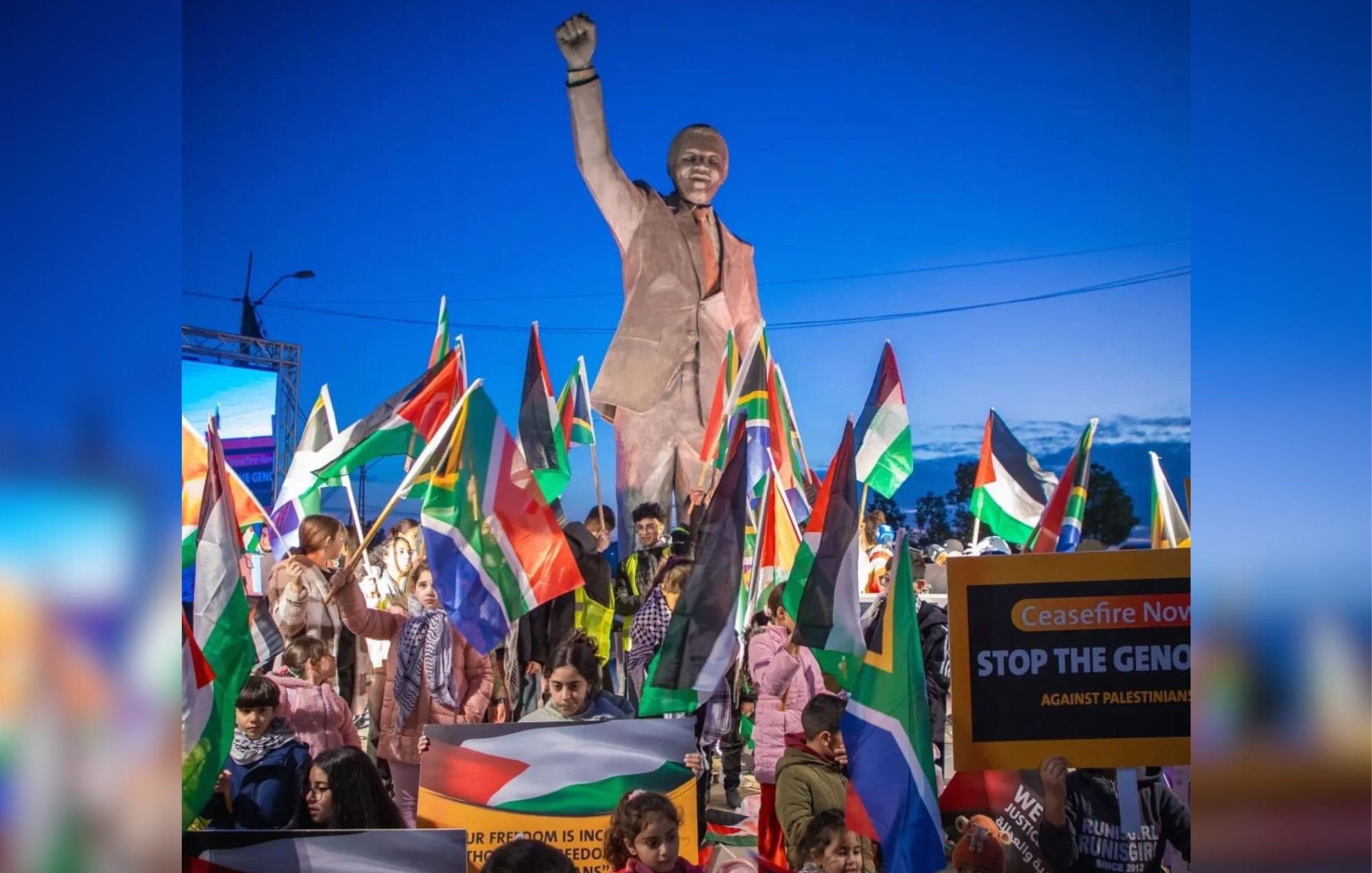 L'image montre une scène de manifestation en plein air, sous un ciel bleu. Au centre, se dresse une grande statue de Nelson Mandela, levant un poing en signe de force et de détermination. Autour de la statue, des manifestants tiennent une multitude de drapeaux, notamment des drapeaux sud-africains et palestiniens, créant un mélange de couleurs vives. Des pancartes sont visibles, certaines appelant à un cessez-le-feu et dénonçant des injustices. Les visages des participants expriment à la fois la passion et l'engagement pour leur cause. L'atmosphère générale est celle de la solidarité et de la lutte pour les droits humains.