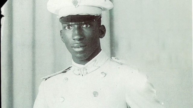 L'image montre un jeune homme en tenue militaire. Il porte un uniforme blanc qui se compose d'une chemise et d'un pantalon assortis, avec une ceinture qui souligne sa taille. Sur sa tête, il a une casquette blanche ornée d'un insigne. Son expression est sérieuse et déterminée. L'arrière-plan est neutre, probablement un drap ou un fond uni, ce qui met en valeur sa silhouette et son uniforme.