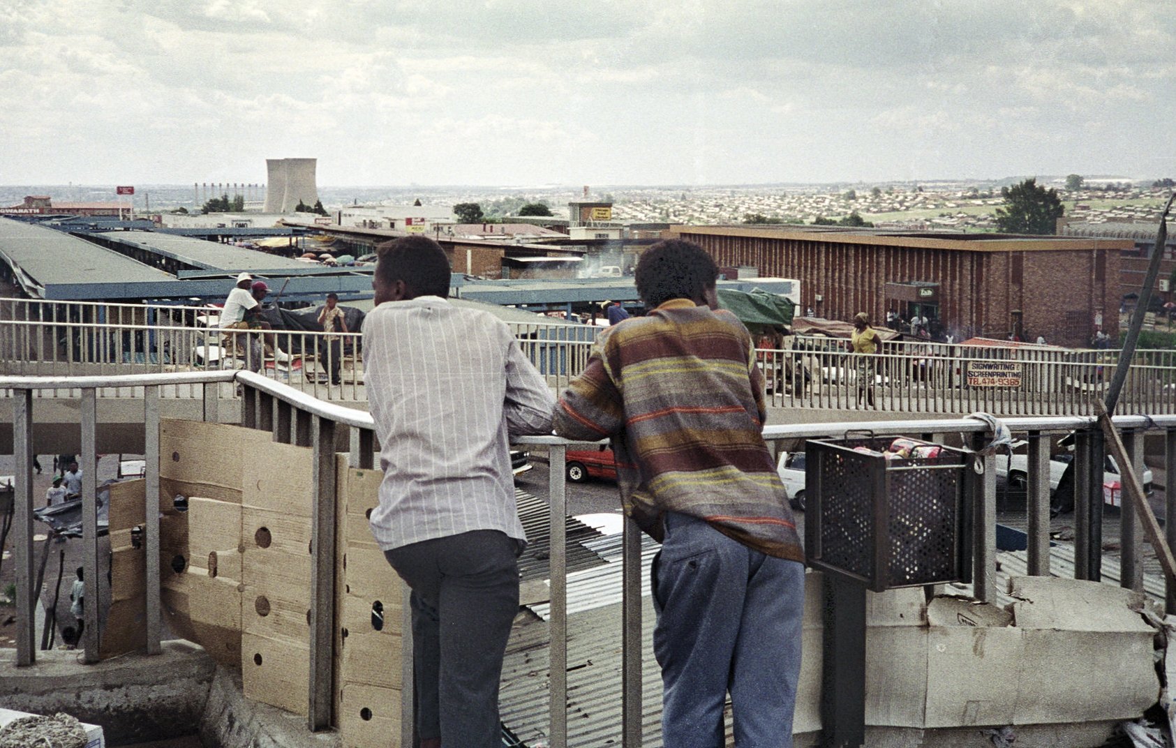 Dans cette image, on voit deux hommes debout sur un balcon surélevé, se penchant contre la balustrade. Ils regardent en bas un marché animé. Leurs silhouettes sont tournées vers l'extérieur, révélant un paysage urbain qui s'étend derrière eux, avec des bâtiments et des petites boutiques. L'environnement est vivant, rempli de personnes et d'activités. Le ciel est nuageux, ajoutant une ambiance mélancolique à la scène. Les vêtements des hommes suggèrent une certaine simplicité, avec des motifs discrets. L'ensemble dégage une atmosphère de contemplation et d'observation de la vie quotidienne en ville.