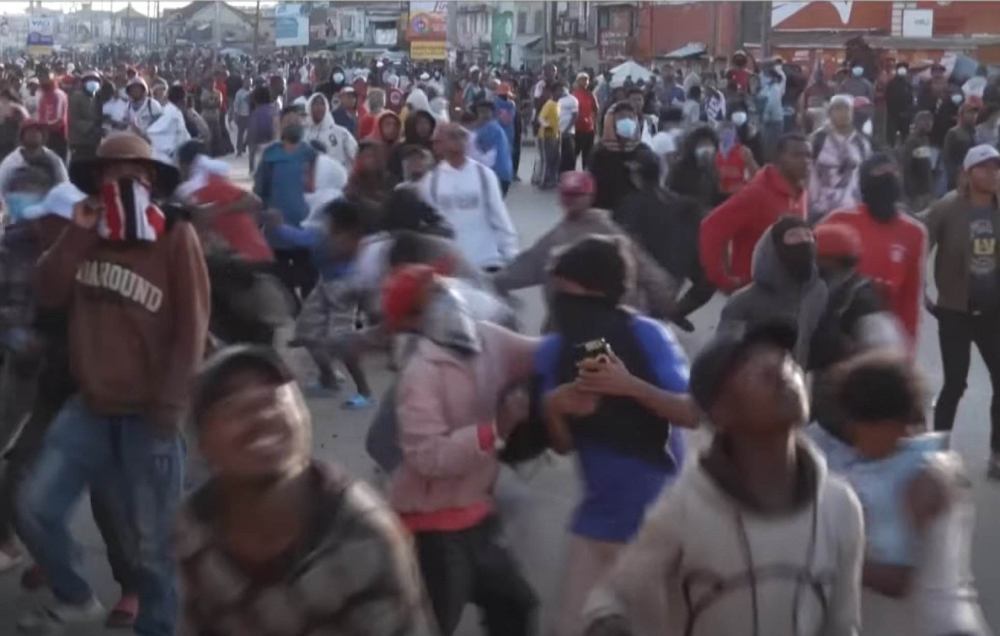 L'image montre une foule dense de personnes rassemblées dans une rue, probablement lors d'une manifestation ou d'un événement populaire. Les participants portent des vêtements variés et beaucoup d'entre eux ont des masques ou des foulards sur le visage. On peut sentir une ambiance animée et énergique, avec des mouvements rapides et des gestes expressifs. Certains individus semblent prendre des photos avec leurs téléphones. L'arrière-plan est rempli d'autres personnes, créant une atmosphère vibrante et collective.