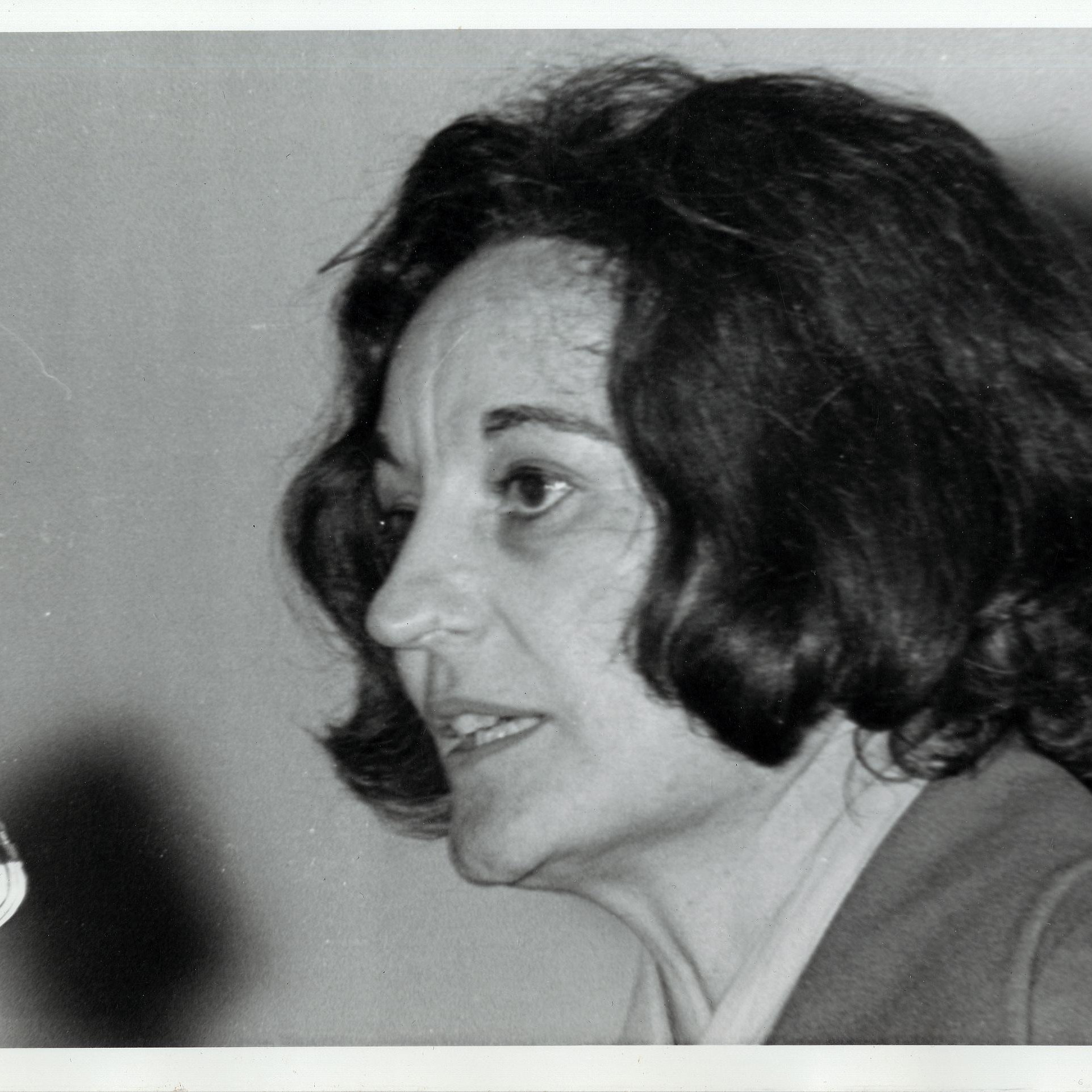 L'image montre une femme avec des cheveux foncés, mi-longs et légèrement ondulés. Elle est en profil, parlant près d'un microphone. Son expression est concentrée, et on peut deviner une certaine passion dans son discours. Elle porte une veste simple, et l'arrière-plan est flou, ce qui met l'accent sur son visage et son expression. L'éclairage est doux, soulignant les traits de son visage.